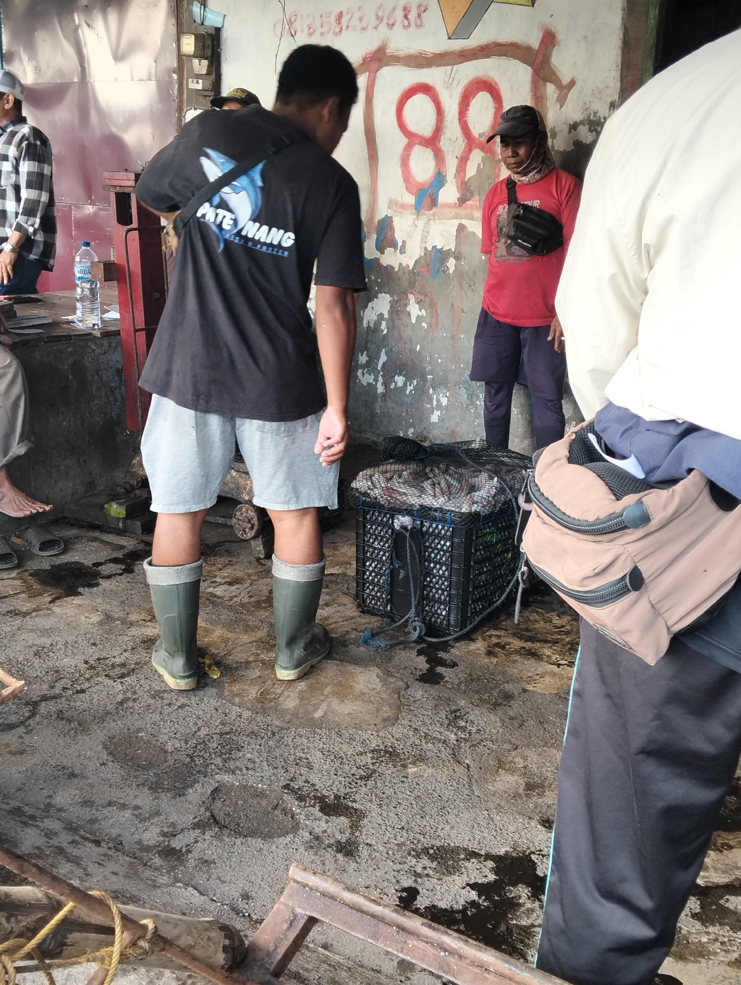 A low-angle shot captures a scene in Muncar, Indonesia, likely a fish market, with three individuals present. In the foreground, a person wearing dark green rubber boots and light grey shorts stands with their back to the camera. To their right, a black plastic crate is filled with fish and covered with a black net. A light brown shoulder bag with zippers is held by a person whose lower body is visible on the far right, wearing black pants with a light blue stripe. In the middle ground, a person in a red t-shirt and dark shorts is visible, wearing a black cap and a black fanny pack. To the left, a person in a plaid shirt is partially visible. The ground is wet and appears to be concrete, with puddles and debris. The background shows a weathered wall with faded graffiti, including red numbers that appear to be "88". A bottle of "AQUA" water sits on a table in the left portion of the image.