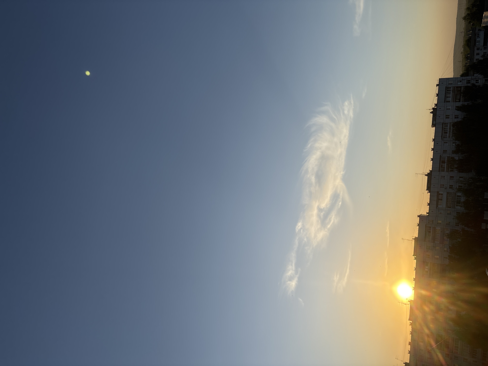 The image shows a horizontal view of a city at sunset. The sky is a gradient of blue to yellow, with wispy clouds near the center. A bright sun is setting behind buildings. On the right, high-rise apartment buildings dominate the skyline. There are trees and other greenery interspersed among the buildings. A bright dot appears in the upper left of the image. The image appears to have been taken in Chaikovsky, Russia.