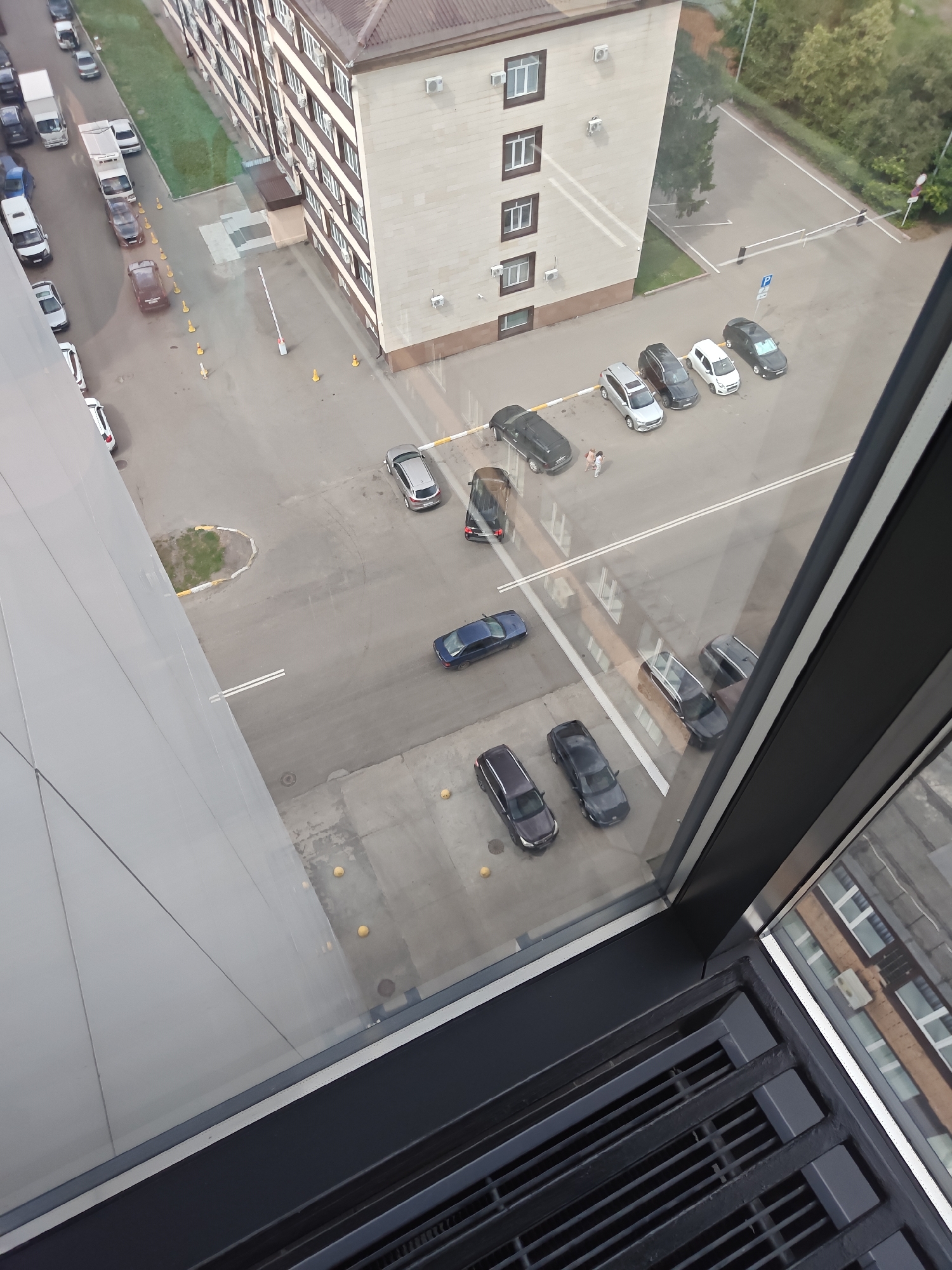 This is a high-angle, outdoor, daytime photograph taken from a window, looking down at a parking lot and a modern, multi-story building with a light-colored facade and brown accents. The building has numerous windows and what appear to be air conditioning units mounted on its exterior. Adjacent to the building is a paved area that serves as a parking lot. Several cars are parked in the lot, including a dark blue sedan, a silver hatchback, several dark-colored SUVs, and a white sedan. Some of the vehicles are on marked parking spaces, while others are parked in a more informal manner. In the foreground, part of the window frame and a ventilation grate are visible. The street area has yellow traffic cones and a retractable barrier. The context of Petropavl, Kazakhstan, suggests an urban or semi-urban environment.