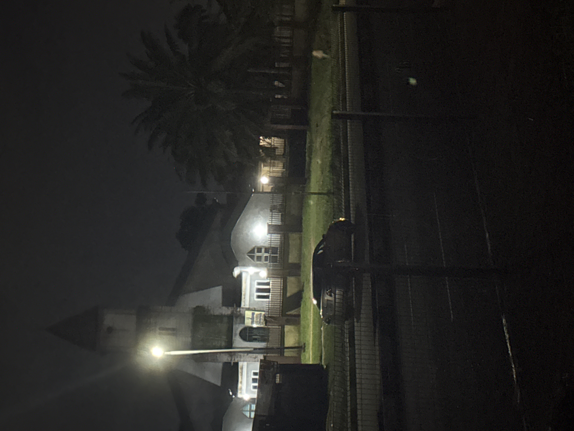 The image depicts a nighttime scene in Afaha Ibesikpo, Nigeria, featuring a building with a prominent tower, possibly a church, illuminated by a streetlamp. To the right of the building, several palm trees are visible against the dark sky. In the foreground, a dark-colored sedan is parked on the side of a road, with a fence separating the road from the building's grounds. The environment appears to be a residential or institutional area, with manicured grass and further fencing around the property. The lighting conditions are low, with the streetlamp and building lights providing the primary illumination, casting long shadows and creating a slightly hazy atmosphere.