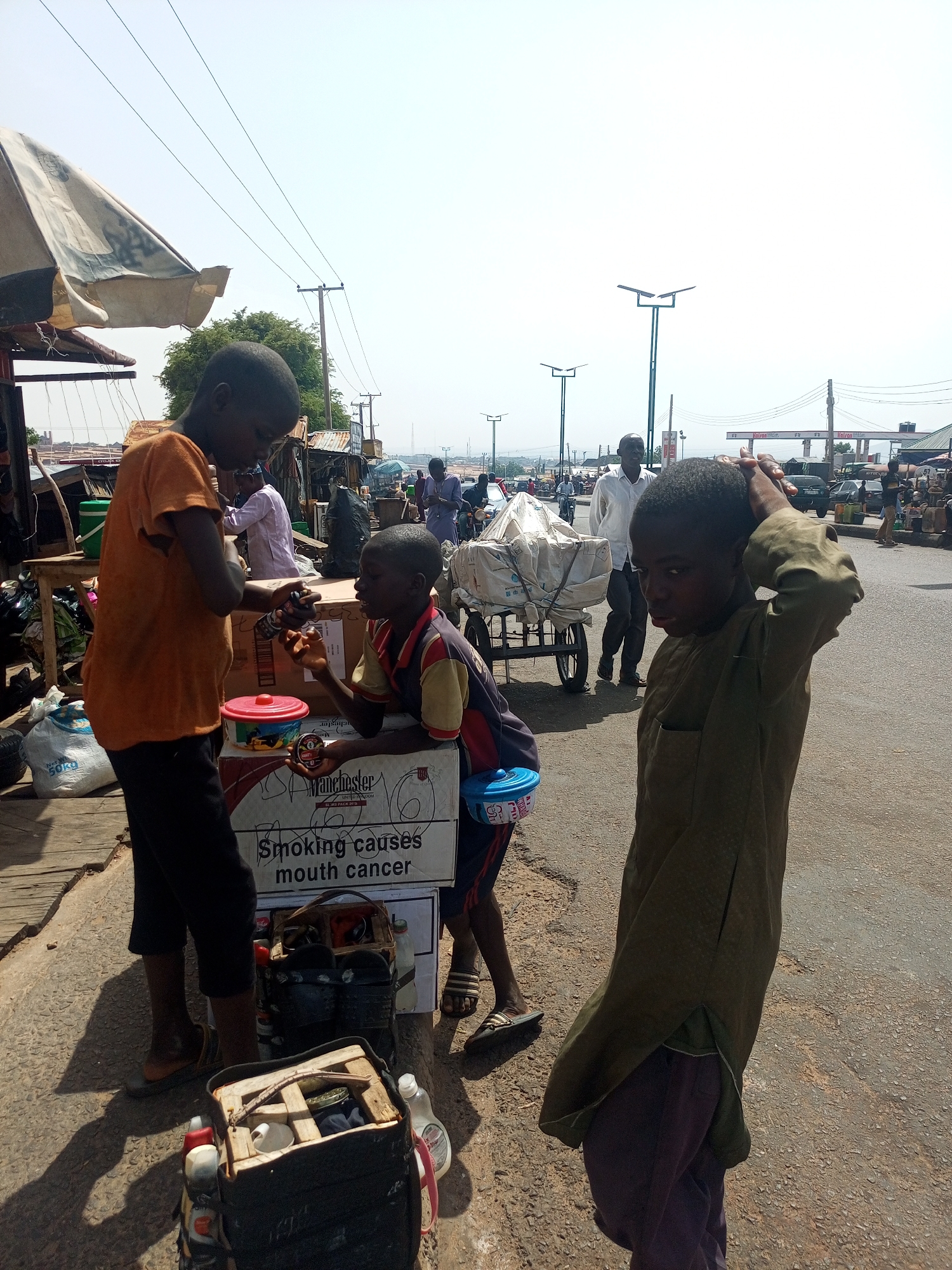 Three boys are positioned in a street scene in the city of Gombe, Nigeria. One boy wears an orange shirt and black shorts and stands near a stack of boxes that say, "Smoking causes mouth cancer." Another boy leans on the same boxes, wearing a multi-colored shirt and sandals. A third boy, in a green shirt and purple pants, stands in the street with his hand on his head. The scene includes other people, a cart loaded with covered items, a street, a sidewalk, and various objects. A covered structure is partially visible. Beyond the immediate group, people and vehicles are seen in the background.