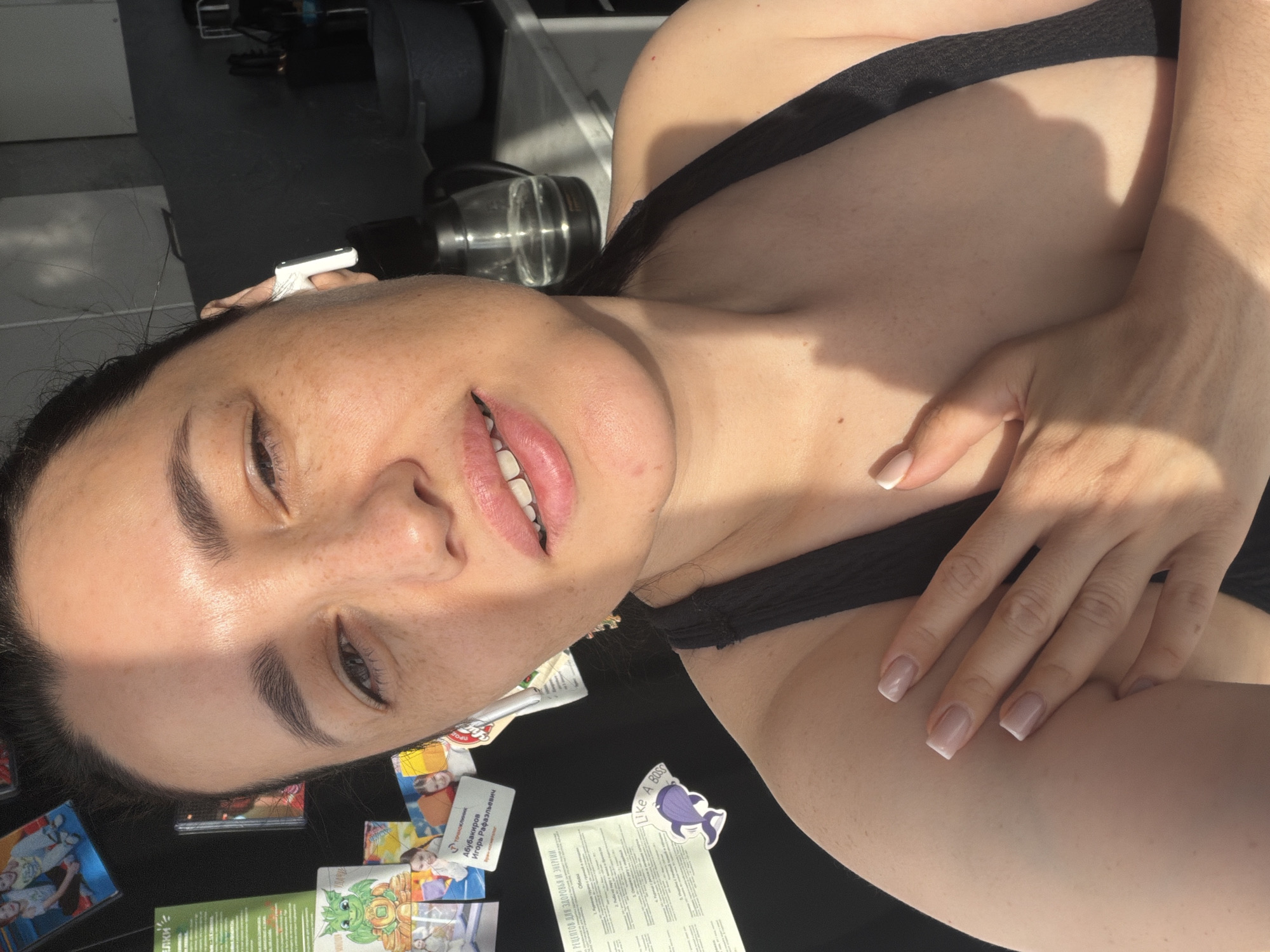 A woman, appearing to be in her 30s, is captured in a close-up, selfie-style photograph. She has light skin with freckles, dark hair pulled back, and is wearing a black tank top. Her left hand rests on her upper chest, and her right hand is partially visible. She has a white earbud in her right ear. The background suggests a domestic setting, possibly a kitchen, with cabinetry and a kettle visible. Several magnets and notes are attached to a dark surface on the left side of the image. The lighting indicates a sunny environment, with harsh shadows cast across her face and body. The image was taken in Mytishchi, Russia.