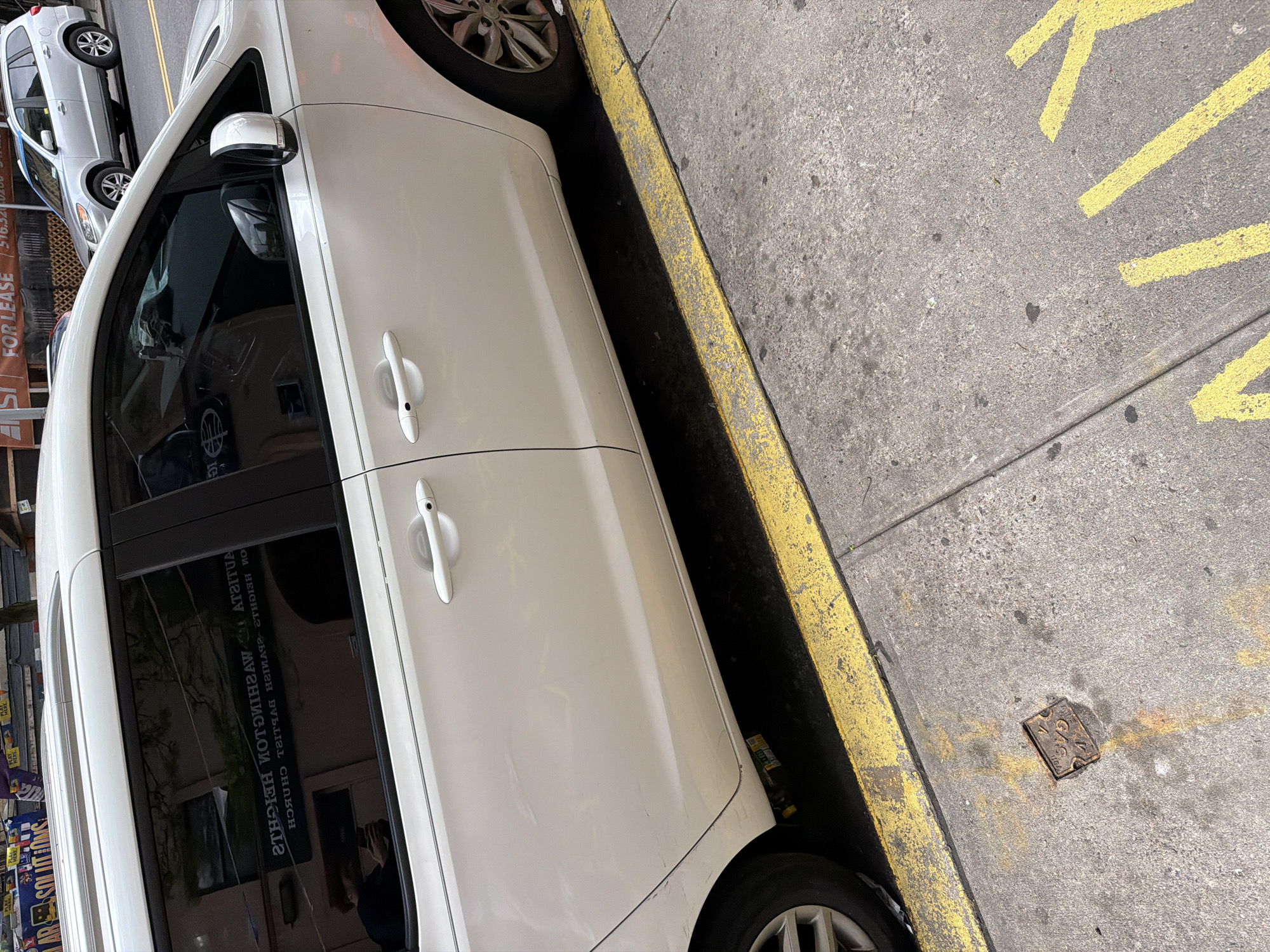 A white minivan is parked on an urban street next to a yellow-painted curb and concrete sidewalk. The image is taken from a perspective looking down at the vehicle's passenger side. The front passenger and sliding rear passenger doors are visible, with their white handles. Reflections in the tinted windows of the minivan show inverted text, including "STRAIGHTAWAY CHURCH" and "CHURCH OF WASHINGTON HEIGHTS," along with a person holding a phone, presumably the photographer.
The concrete sidewalk features yellow painted markings, which include a visible "K" and what appear to be parts of arrows or other letters, suggesting a designated area. A small, square metal plate, possibly a utility marker, is embedded in the sidewalk with the inscription "5 / G / 6 / N.Y.C." and other faint symbols.
In the background, another vehicle, a silver SUV or minivan, is parked behind the white minivan. Further in the background, a building facade displays a brown sign with "FOR LEASE" written in white, along with a partial phone number starting with "516.52". Other signs, including one with the word "SOLUTIONS," are partially visible but mostly illegible. The scene is illuminated by daylight, suggesting clear weather conditions in an urban setting, consistent with New York, USA.