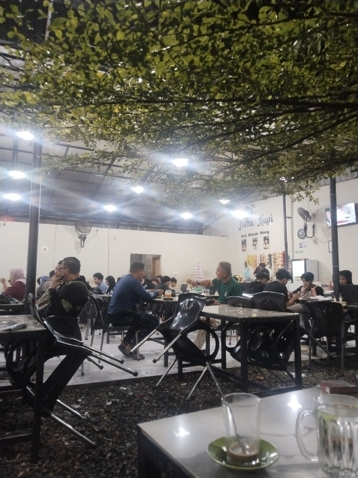 The image captures an evening scene in a cafe located in Lhokseumawe, Indonesia. Several people are seated at tables, engaged in conversations or activities such as eating and drinking. The cafe has an open-air design with a roof structure adorned with artificial foliage and illuminated by overhead lights. Tables and chairs are arranged throughout the gravel-covered area. A sign with the name "Doha Kupi" is visible in the background, along with a television screen mounted on the wall. Drinkware is present on the tables in the foreground.