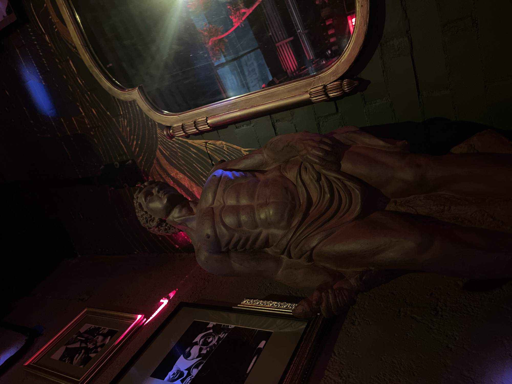 An indoor photograph shows a statue of a nude male reclining, with two framed photos underneath it, lit with red and blue lights. Above the statue, a large, ornate gold mirror reflects the scene. The location is most likely the bar Gin Gin, in Puebla, Mexico, known for its dramatic decor.