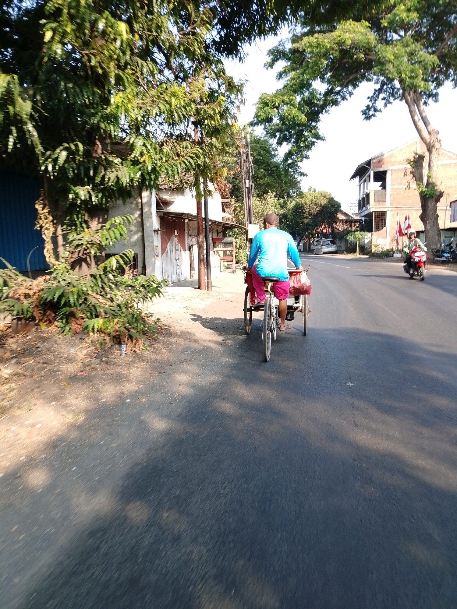 A man wearing a bright blue long-sleeved shirt and pink shorts rides a three-wheeled bicycle with a trailer on a paved road in Pasuruan, Indonesia. The bicycle is being pedaled from behind, with the trailer carrying a red bag. The man is wearing sandals. In the background, there are buildings, trees, and other vehicles on the road, including a red motorcycle carrying a person with a white helmet. The scene appears to be in a semi-urban area with a mix of residential and commercial structures. The road is asphalt, and there are shadows from trees cast on the road. The sky is clear and bright, suggesting it is daytime.
