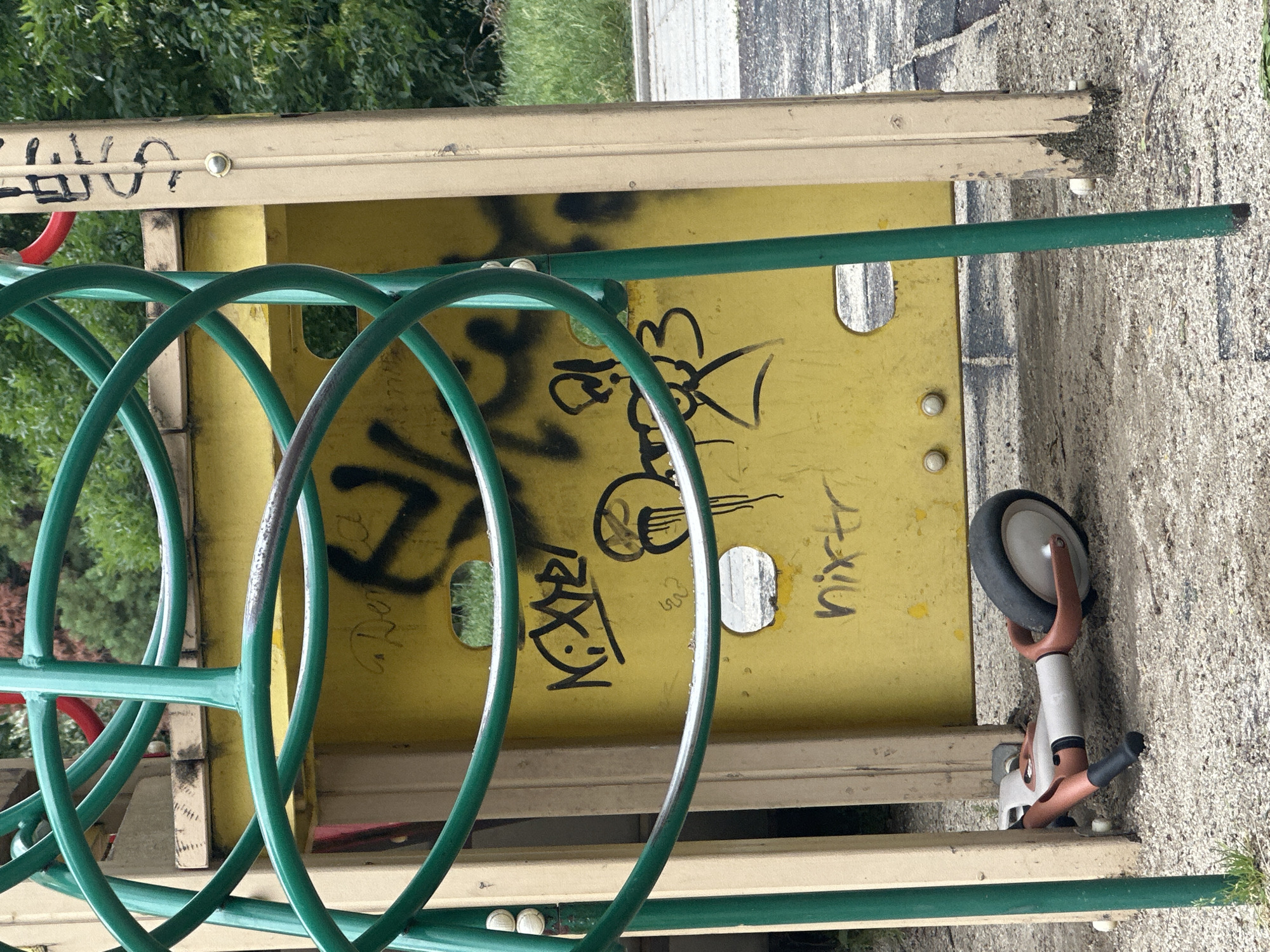 The image shows a playground structure with green metal rings and a yellow panel covered in graffiti. A small, silver and orange scooter is lying on the ground in front of the structure. The ground is covered in gravel and some dirt. In the background, there are trees and a paved area. The graffiti includes words and drawings. The overall impression is that of a neglected and vandalized playground. The image does not provide any explicit details about the location in the city of Sofia, Bulgaria, but the presence of a playground suggests an outdoor public space.