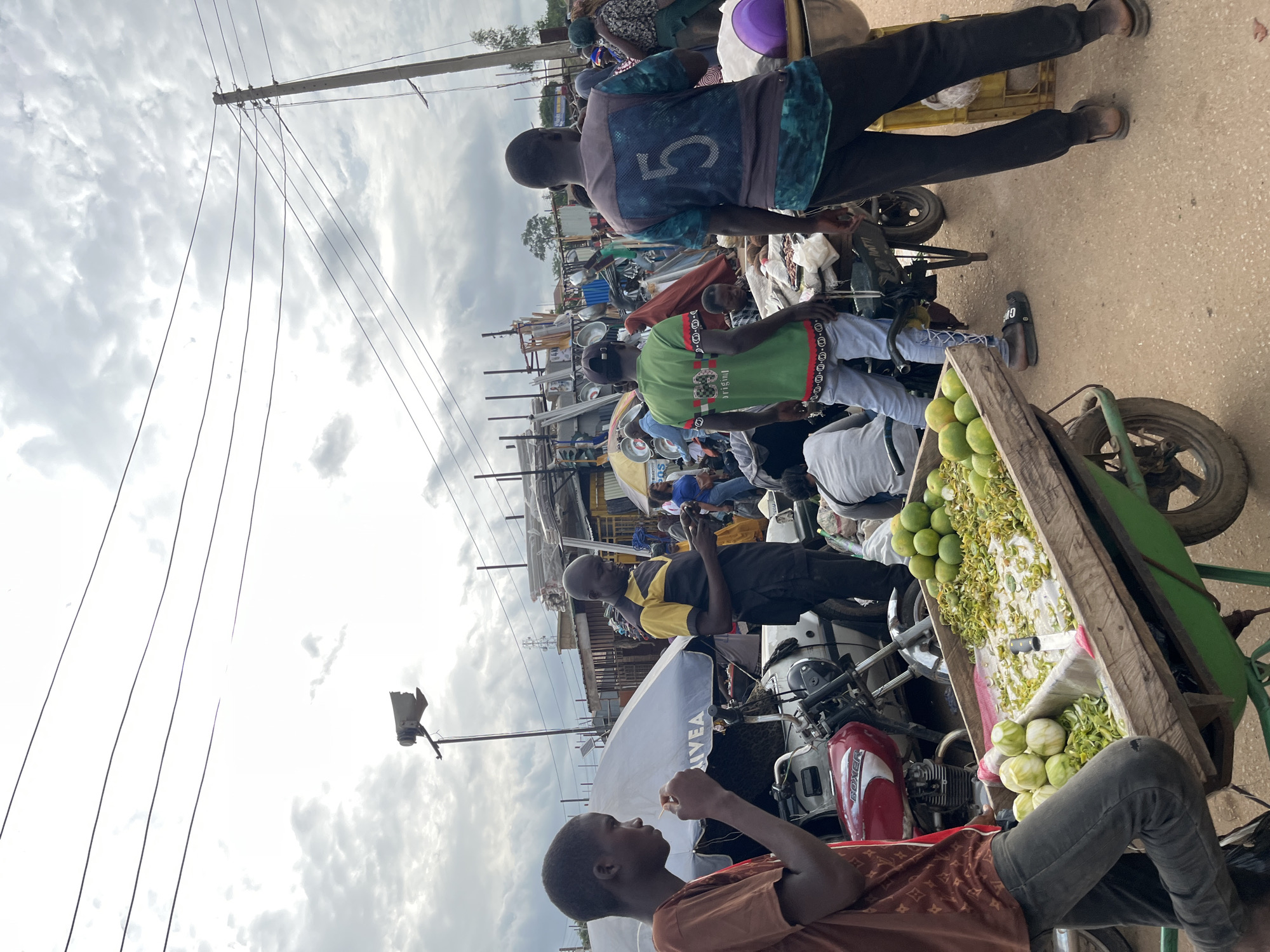 This image captures a bustling outdoor market scene in Gura Top, Nigeria. The sun is obscured by a cloudy sky, suggesting it's likely daytime. The foreground is dominated by people and various objects, including several motorcycles, some of which appear to be modified for carrying goods. A wooden crate overflowing with green citrus fruits, possibly limes or lemons, is prominently displayed on one of the motorized carts. Alongside the fruits, there are bundles of what appear to be dried herbs or spices.
Several individuals are visible, engaged in various activities. One person in the foreground, wearing a brown patterned shirt and dark pants, is sitting and looking to their left. Behind them, a person in a green shirt with a black and white patterned trim is standing. Further back, another individual in a blue and black patterned vest with the number "5" on it is also standing. The overall atmosphere suggests a lively marketplace with people gathering and potentially trading goods.
The presence of power lines strung across poles indicates an urban or semi-urban setting. Some of the motorcycles have visible branding, such as "BOXER" on the fuel tank of a red motorcycle. The ground appears to be a paved surface, possibly asphalt or concrete, with visible speckled patterns. The general impression is one of everyday commerce and activity within a community.