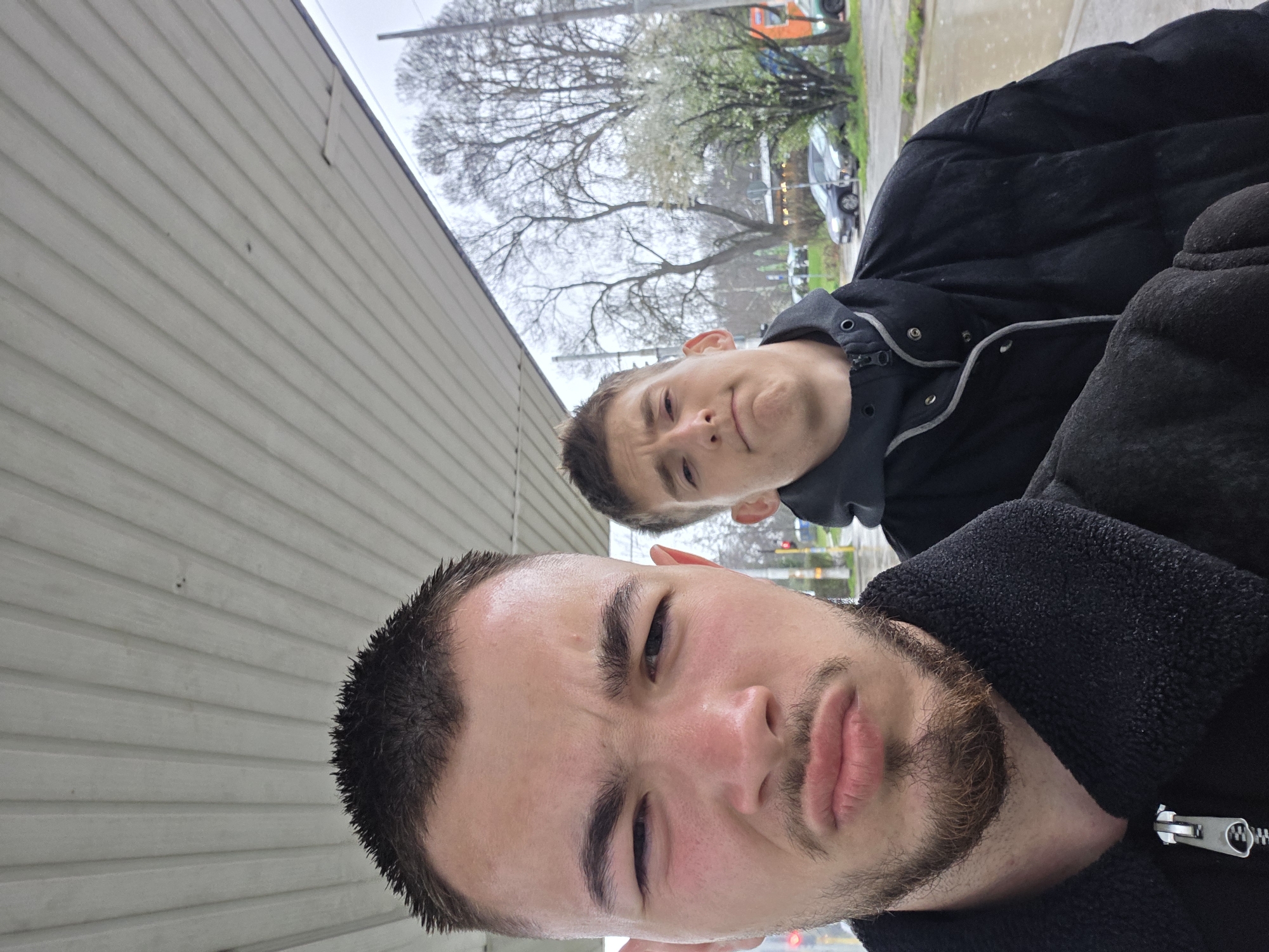 Two young men are posing for a selfie outdoors. The man in the foreground has a dark beard and short dark hair. He is wearing a black jacket. The second man is behind him, slightly out of focus, with short brown hair. He is also wearing a black jacket with a hood. In the background is a building with gray horizontal siding and bare trees. Cars and streetlights are visible further in the background. The location is likely an urban or suburban area. No information is provided to indicate this image was taken in Sofia, Bulgaria.