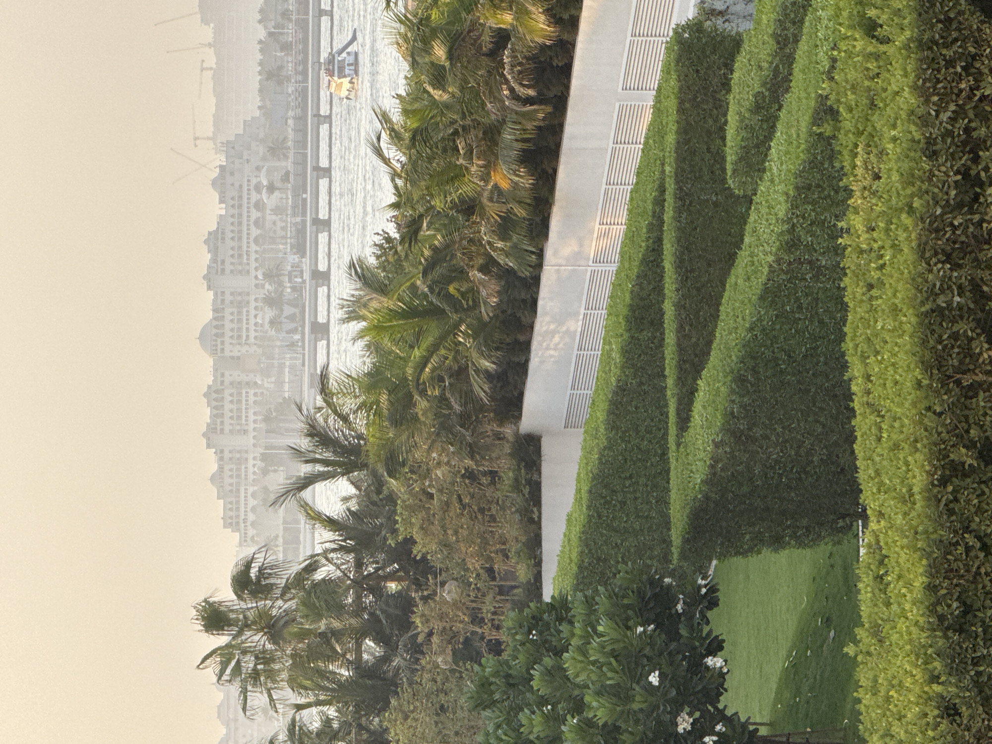 This image captures a serene view of the Dubai coastline, likely from a high vantage point. In the foreground, meticulously trimmed hedges form a geometric pattern, leading the eye towards a lush green lawn with a tree adorned with white flowers. Beyond this manicured garden, a body of water shimmers, with a small boat visible in the distance. Palm trees frame the scene, their fronds swaying gently. In the background, the distinctive silhouette of Dubai's modern architecture emerges from a hazy sky, suggesting a warm, possibly humid, day. The architecture appears grand and ornate, characteristic of luxury hotels or resorts. Construction cranes are visible in the far distance, indicating ongoing development. The overall impression is one of tranquility and upscale leisure.