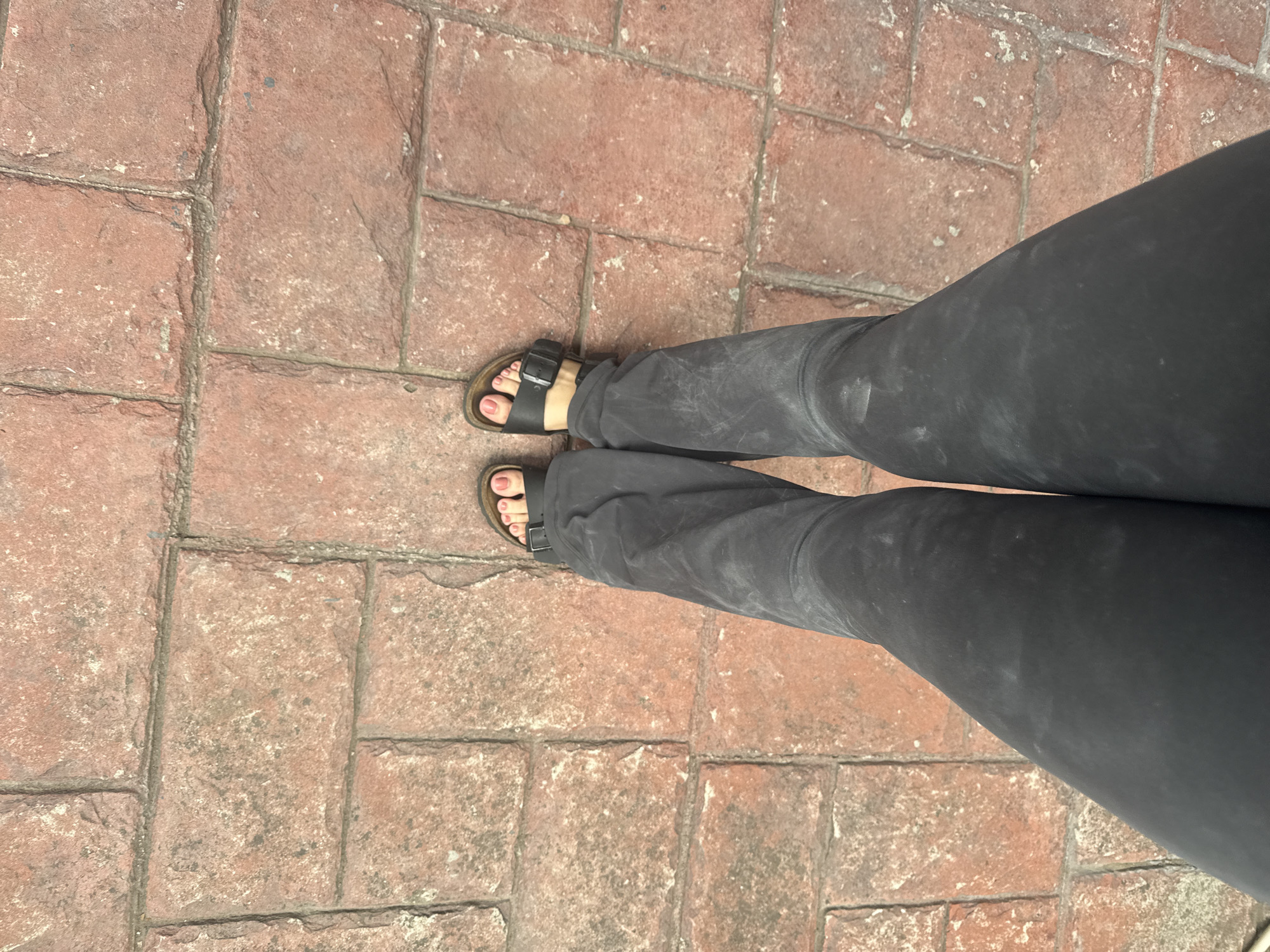 The image shows a top-down view of a person's legs and feet standing on a brick-paved surface. The person is wearing dark-colored pants and black Birkenstock-style sandals, with pink nail polish visible on their toes. The brick pavement consists of rectangular bricks arranged in a pattern. The overall impression is a casual, everyday scene. Given the additional location information, the scene is situated in San Andrés Cholula, Mexico.