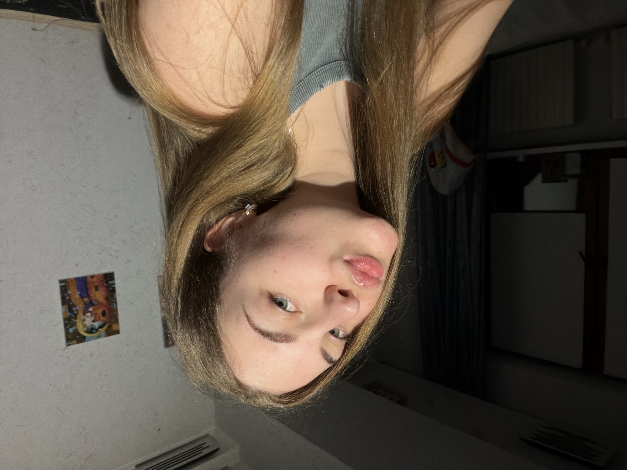 A young woman with long blonde hair and blue eyes is pictured in a selfie. She is wearing a grey tank top and a delicate necklace. Her lips are slightly pursed, and she is looking towards the camera. A small, square earring is visible in her right ear. In the background, a wall with a textured pattern is visible, with two framed pictures hanging on it. To the left, a window with dark curtains and a radiator can be seen. An air conditioning unit is mounted high on the wall. The lighting suggests an indoor setting, likely an apartment in Dnipro, Ukraine, given the context.