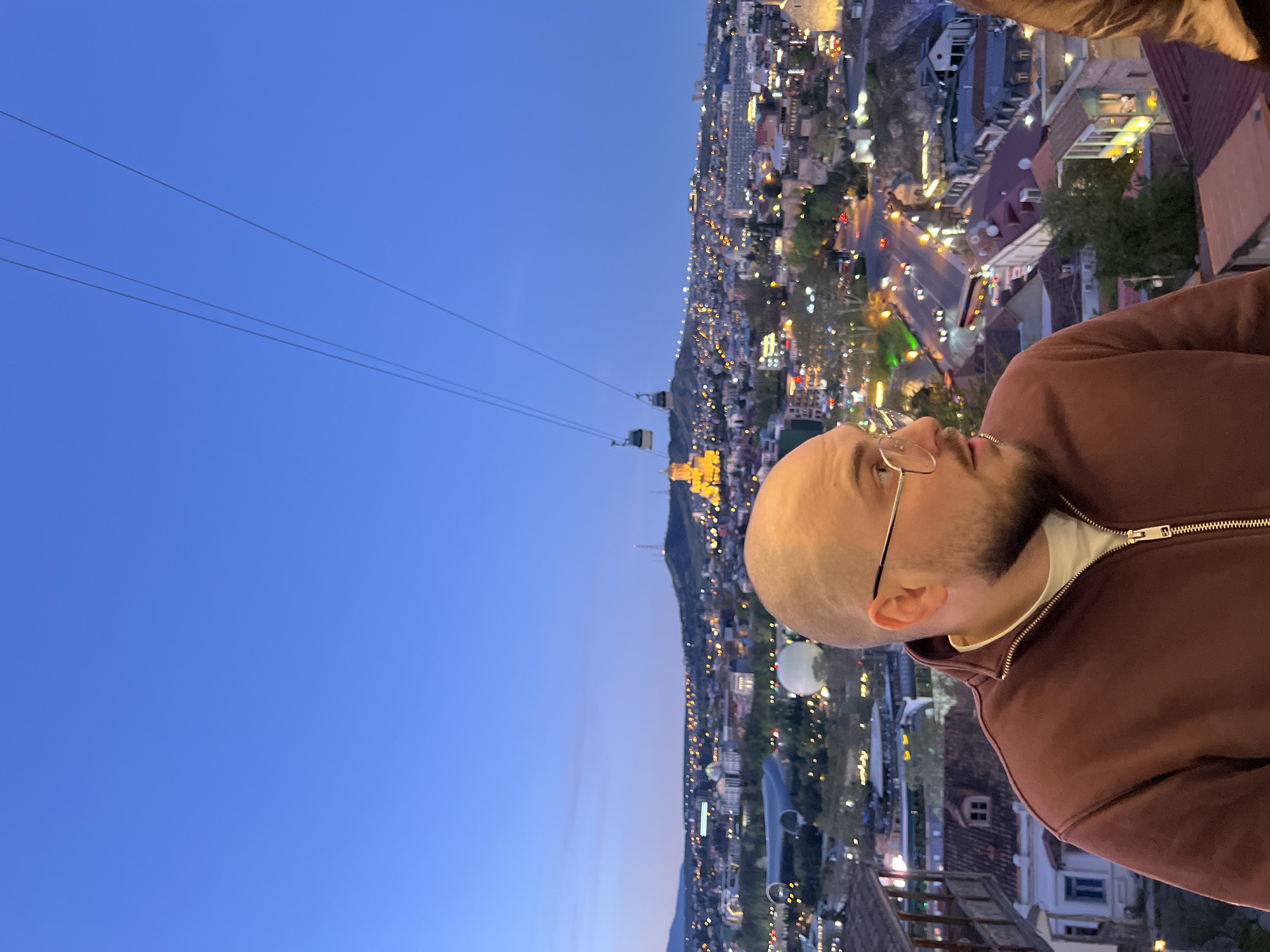 A medium shot depicts a man with a shaved head, wearing glasses and a maroon zip-up jacket over a white undershirt, gazing toward the left. The background reveals an urban landscape with numerous lights, suggesting a city at dusk or night. Two cable cars are suspended over the city, indicating a location with mountainous terrain or significant elevation changes. Based on the architectural style and presence of the cable cars, the image is likely taken in Tbilisi, Georgia.