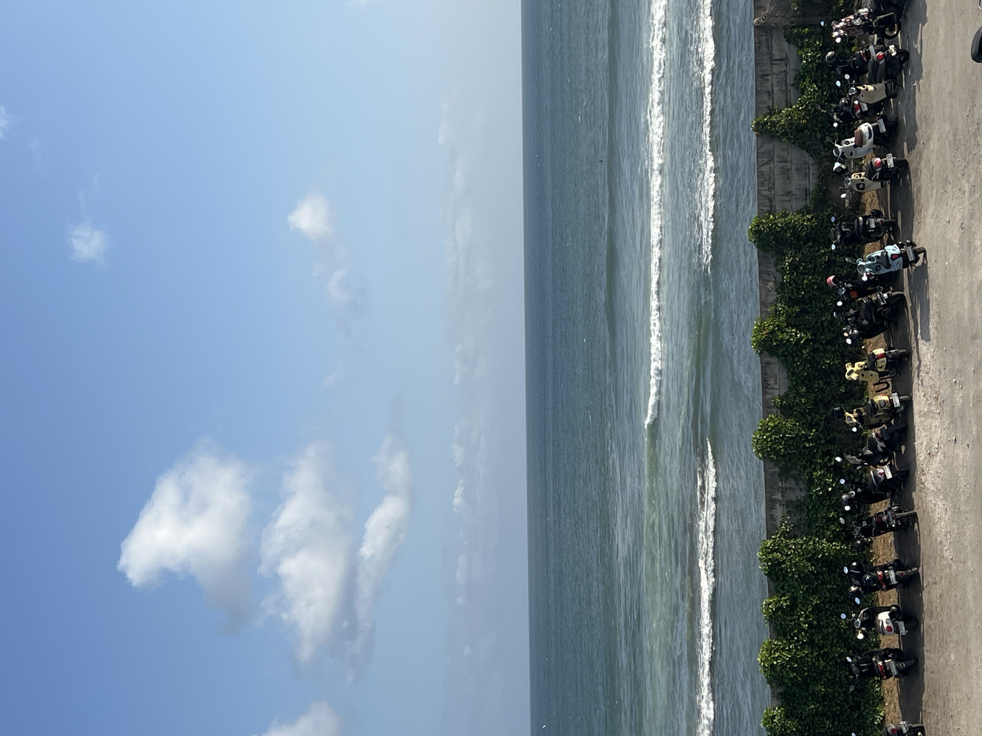 A serene view of the ocean is captured from a slightly elevated vantage point in Mengwi, Indonesia, on a bright, clear day. The sky is a vibrant blue, dotted with fluffy white clouds. The vast expanse of the ocean stretches to the horizon, with gentle waves rolling towards the shore. A concrete wall, partially covered with lush green ivy, separates a parking area from the ocean. The parking area is filled with a variety of parked scooters, mostly dark colored, with a few lighter colored ones interspersed. The ground is a mix of gravel and asphalt. The scene evokes a sense of tranquility and the relaxed atmosphere of a coastal town.