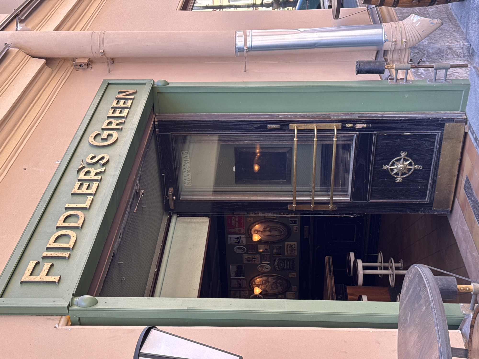 The image depicts the entrance of "Fiddler's Green," an establishment located at Bolshaya Konyushennaya Ulitsa, 1, in Saint Petersburg, Russia. The building's facade is a light peach-pink color, featuring a dark green entrance frame. Above the doorway, "FIDDLER'S GREEN" is spelled out in gold, three-dimensional, slightly weathered letters.
A dark-colored, possibly black, wooden door with a large glass panel on its right side is slightly ajar, opening inward. The glass panel faintly displays "NO SMOKING" from the interior. The door is equipped with brass-toned horizontal pull bars and features a prominent brass-toned compass rose emblem on its lower wooden section.
Through the open doorway, the interior of the pub is visible, showing a bar counter and two white-legged bar stools. The interior walls are decorated with numerous framed pictures and oval wall sconces emitting warm light; one visible frame displays a "Jack Daniel's Whiskey" advertisement.
To the left of the entrance, a partial view of a white, rectangular light fixture is visible. On the right side of the entrance, a silver-colored metal downspout or ventilation pipe is vertically attached to the building wall with metal bands. Below this pipe, a smaller, discolored pipe leads into a gutter structure above a stone block base. A slender, dark-colored metal stand with horizontal bars and a cylindrical top is affixed to the right side of the green door frame. Partially visible on both the left and right sides of the foreground are dark-colored round tables with barrel bases. No people are present in the scene. The lighting suggests daytime.
