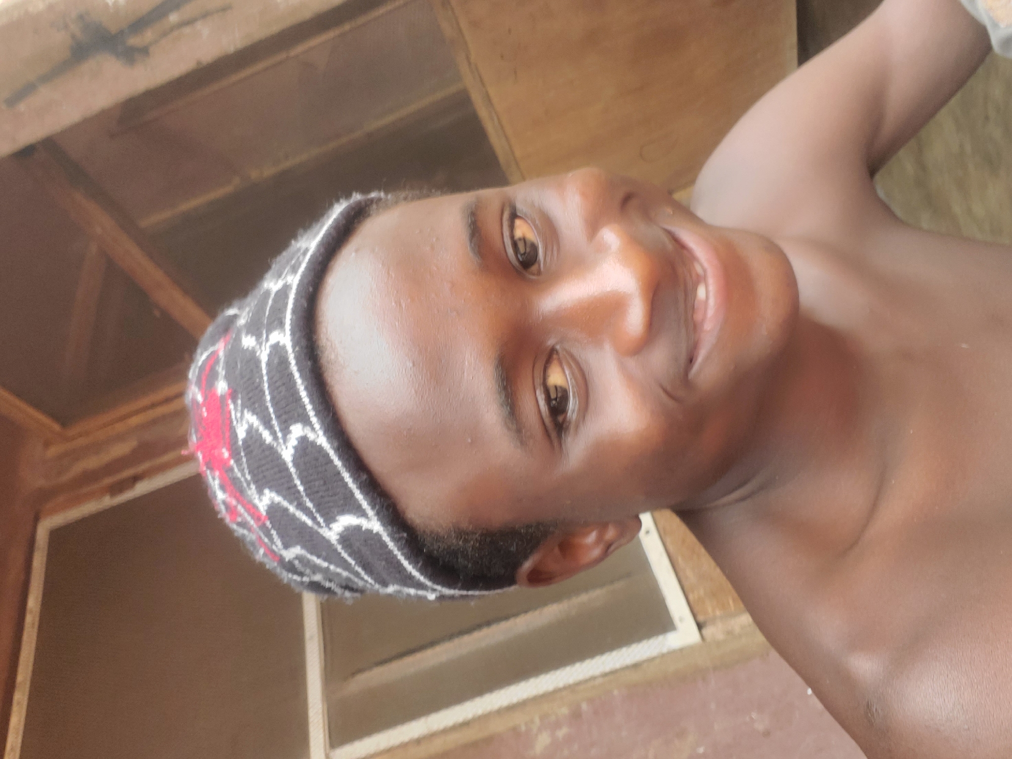 A young, dark-skinned man is depicted from a low-angle perspective, smiling and looking toward the viewer. He is wearing a knitted cap with a black and white geometric pattern and a red design. The man is shirtless. The setting appears to be indoors, near a window with a visible frame. Wooden elements are present in the background. There is no definitive information in the image that confirms it was taken in the city of Onitsha, Nigeria. The description would need to be based on the visible image only.