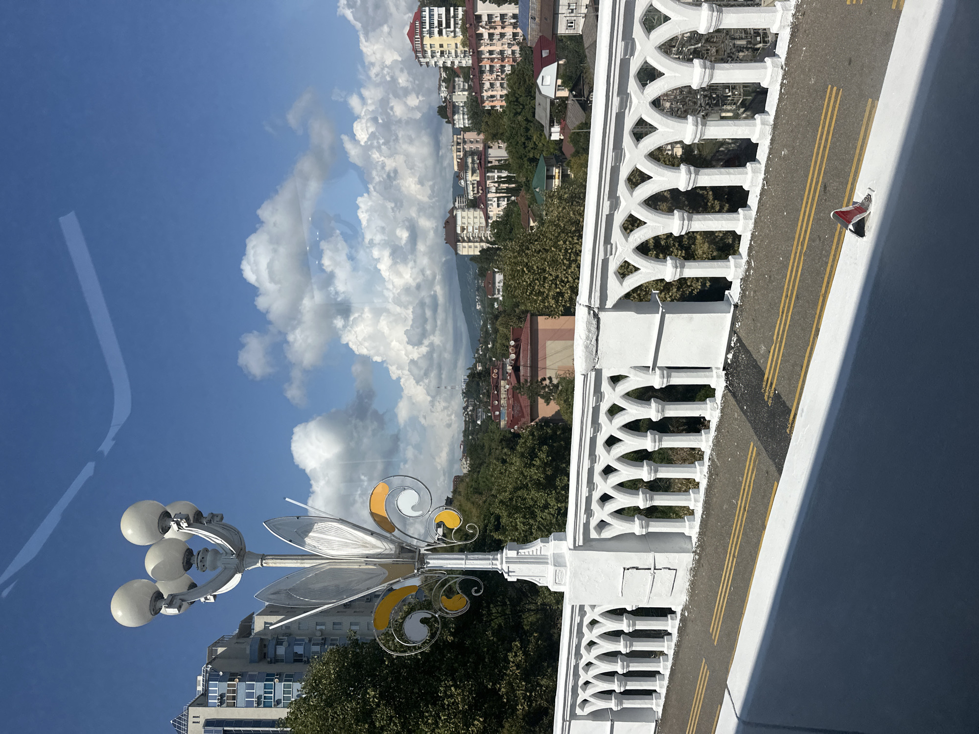 The image depicts a street scene in Sochi, Russia, captured from an elevated perspective, likely a bridge or overpass. In the foreground, a white, ornate balustrade lines the edge of the roadway. The balustrade features a repeating pattern of arched openings supported by columns. A lamppost with a decorative silver finial and four spherical white lights stands prominently to the left of the balustrade. The road surface is asphalt with double yellow lines marking traffic lanes, and a small red and white traffic cone is visible on the road's edge.
Beyond the balustrade, a cityscape unfolds with a dense cluster of buildings, varying in height and architectural style. Many of these are apartment buildings with multiple stories, featuring balconies and numerous windows. Residential houses with pitched roofs are interspersed among them. Lush green trees dominate the mid-ground, softening the urban landscape and indicating a temperate climate. In the background, rolling hills rise, partially obscured by more buildings and foliage, suggesting the hilly terrain characteristic of Sochi. The sky above is a vibrant blue, punctuated by fluffy white cumulus clouds, indicating fair weather. The overall impression is a bright, sunny day in a coastal city known for its mountainous backdrop.