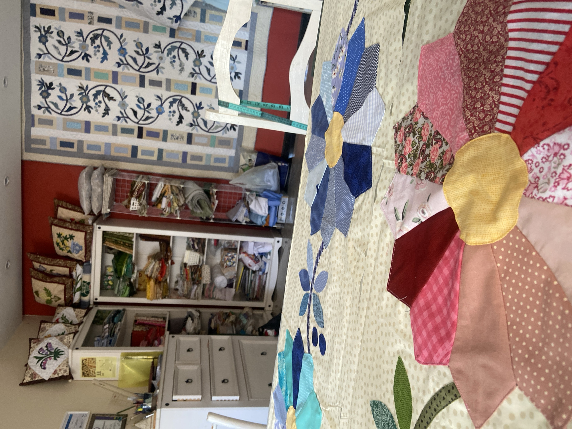A close-up shot focuses on a partially completed quilt laid out on a table. The quilt features a large, stylized flower design with fabric petals in various colors and patterns, including pink gingham, floral prints, solid red, polka dots, and stripes. Another, smaller blue flower motif is visible in the background on the same quilt. The background of the image reveals a room with shelving units filled with fabric and craft supplies, and a large framed quilt hanging on the wall. A white chair with a teal measuring tape draped over it is positioned near the table. The setting appears to be a craft room or sewing studio.