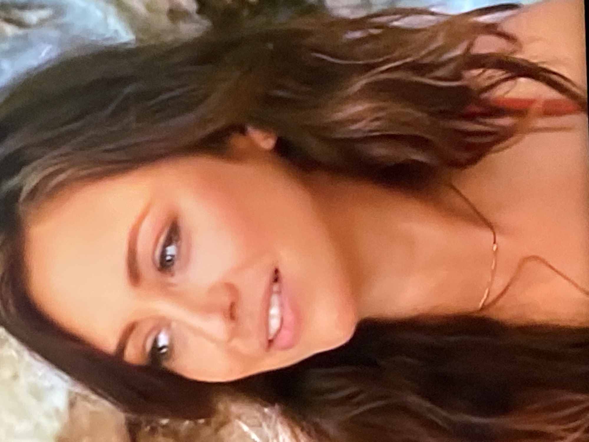 The image shows a close-up, eye-level portrait of a woman from the shoulders up. She has long, dark brown, wavy hair and is wearing a delicate gold necklace. Her skin is warm-toned with subtle blush and light makeup. Her left eye is looking slightly off-camera to the left, while her right eye is looking more directly towards the viewer. Her lips are slightly parted, revealing a hint of teeth. The background is indistinct and appears to be a textured, light-colored surface, possibly a wall or fabric. There are no discernible elements in the image that would allow for the identification of a specific location in the city of Yekaterinburg, Russia, or any other geographical area.