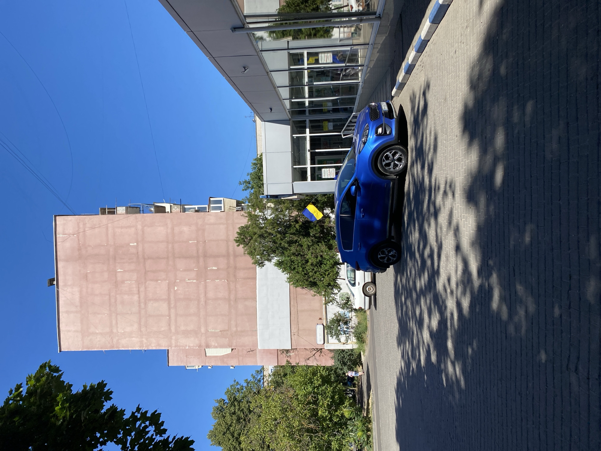 The image captures a bright, sunny day in the city of Chornomorsk, Ukraine. In the foreground, a blue Kia Sportage is parked on a paved area marked with blue and white curbs. The car's license plate is visible, reading "BH 6347 PP". Behind the car, a modern building with large glass windows and a gray facade stands to the right. A Ukrainian flag, yellow and blue, is displayed on a flagpole to the left of this building. To the left of the Kia, a white sedan is parked. Further back, a tall, pink apartment building with a weathered facade dominates the skyline, with a large, light-colored rectangular section on its lower half. Green trees and foliage are present, particularly on the left side of the image, casting dappled shadows on the paved ground. A pedestrian, wearing a light-colored shirt and shorts, is walking away from the camera on a sidewalk near the apartment building. The sky is a clear, vibrant blue.