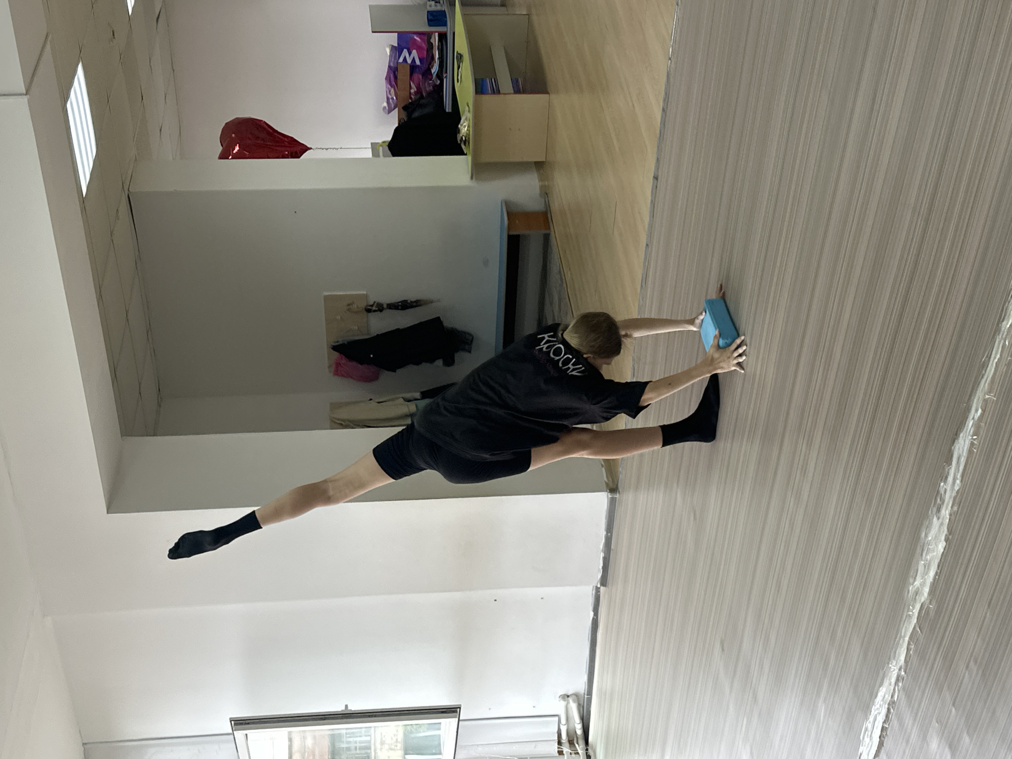 A gymnast in black socks, black shorts, and a black T-shirt with writing on the back is performing a split on a dance studio floor in Votkinsk, Russia. The gymnast is holding a blue block with both hands on a light wood-grained floor. The dance studio has white walls and fluorescent lights. Other objects in the room include a red balloon, storage units, and personal belongings on a shelf. A window is visible.