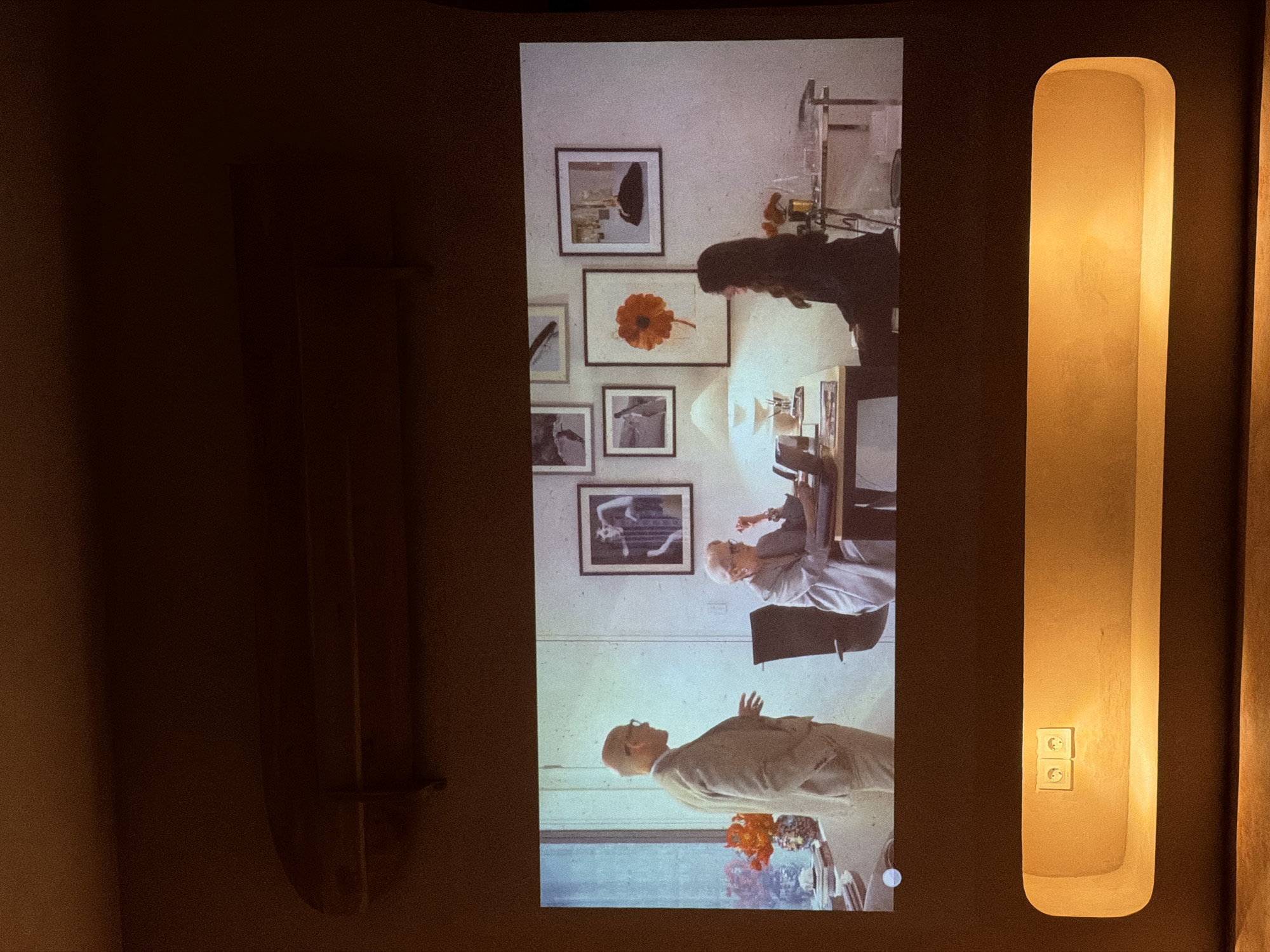 The image depicts a room with a projector screen displaying a scene with two people. One person, possibly a woman, is standing near a desk with a lamp and several framed pictures hanging on the wall. The other person appears to be an older man seated at the desk.
To the right of the screen is a vertical, rounded, recessed light fixture with two electrical outlets. Another similar, but out-of-focus, light fixture is to the right. To the left of the screen is a vertical fixture with one support. The background walls are dark.
The location and city (North Kuta, Indonesia) are not evident from the image.