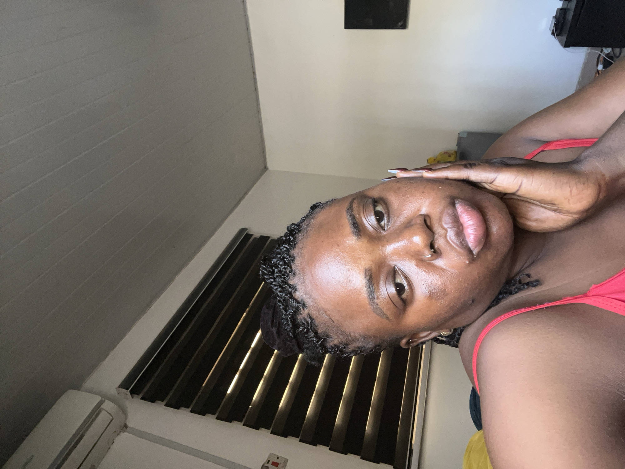 The media file is a portrait of a young Black woman, likely taken in a bedroom in Abuja, Nigeria. The woman is angled with her head tilted downwards, her face partially obscured by her hand. She has dark, braided hair and is wearing a red top. Her skin appears dewy, suggesting it may be morning or that she has recently applied skincare. She has several facial piercings, including one on her nose and another on her lip. The background is dimly lit, with vertical blinds on the right side of the frame casting shadows across the wall. An air conditioning unit is visible on the left. The overall mood appears relaxed and intimate. The image is oriented sideways.
