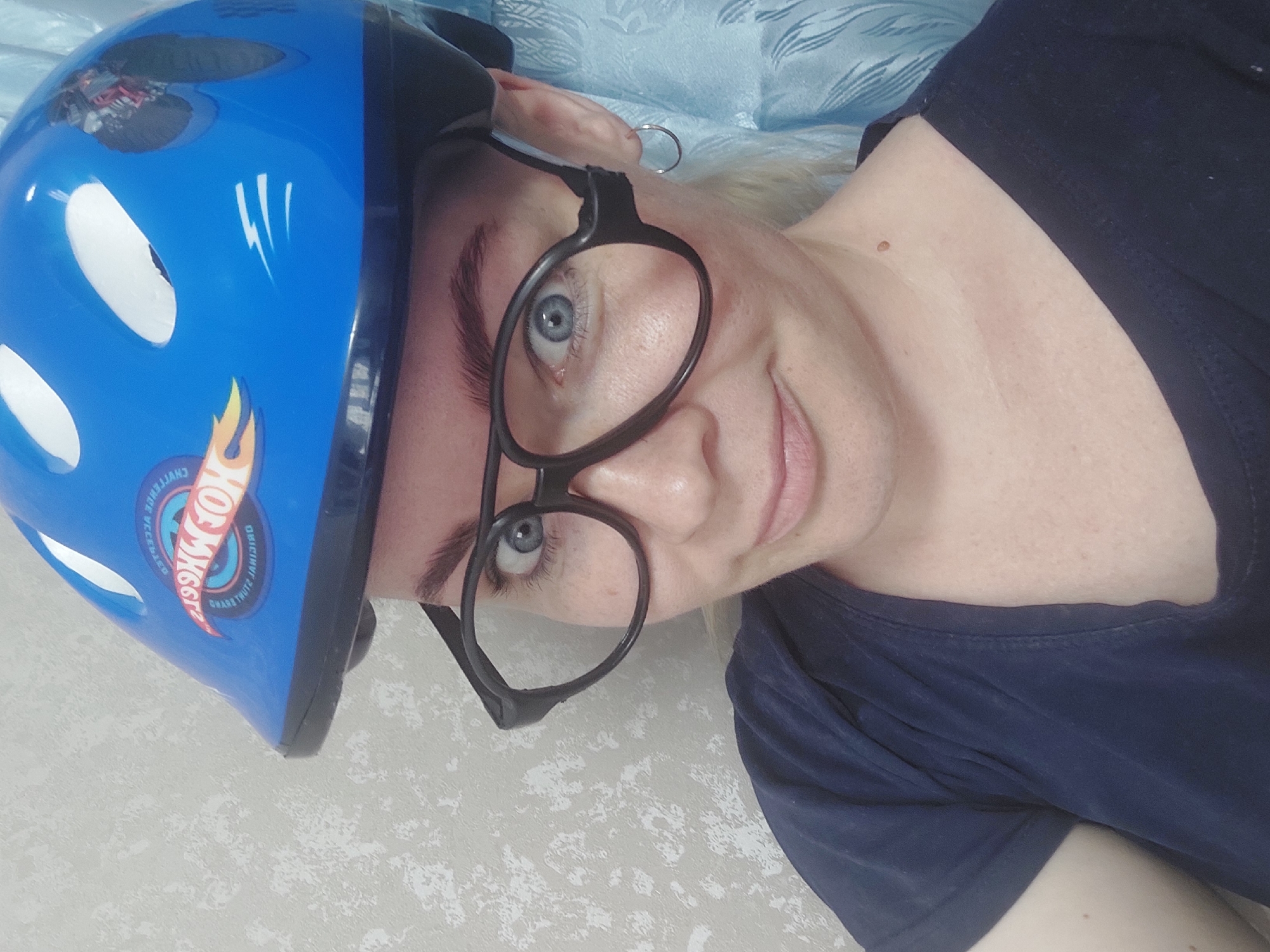 A close-up selfie shows a woman with fair skin, light brown eyebrows, and blue eyes wearing a blue helmet with a "Hot Wheels" logo. She is also wearing large, black-rimmed glasses and a dark blue t-shirt. A silver hoop earring is visible in her right ear. The background is a textured light-colored wall and a blue curtain. The image quality is low, with visible noise and pixelation. There is no indication of the location within the city of Chaikovsky, Russia, or any other specific geographic context. The scene appears to be indoors.