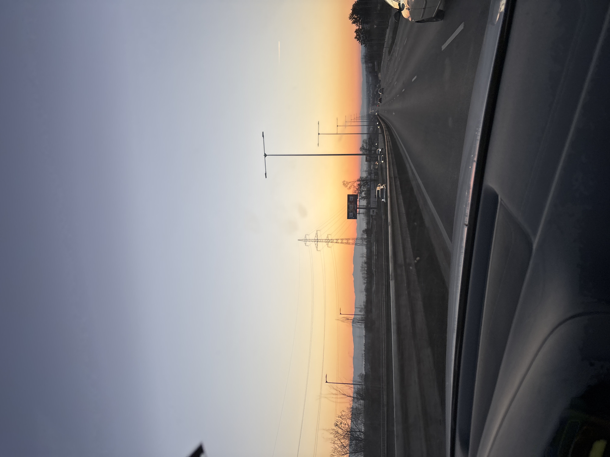 The image shows a highway at dusk, likely taken from inside a moving vehicle. A multi-lane road stretches into the distance, illuminated by streetlights. Power lines run parallel to the highway on the left. The sky transitions from blue to orange towards the horizon. The side of the vehicle is visible in the foreground on the right. The location is possibly near Sofia, Bulgaria, considering the landscape and infrastructure style.