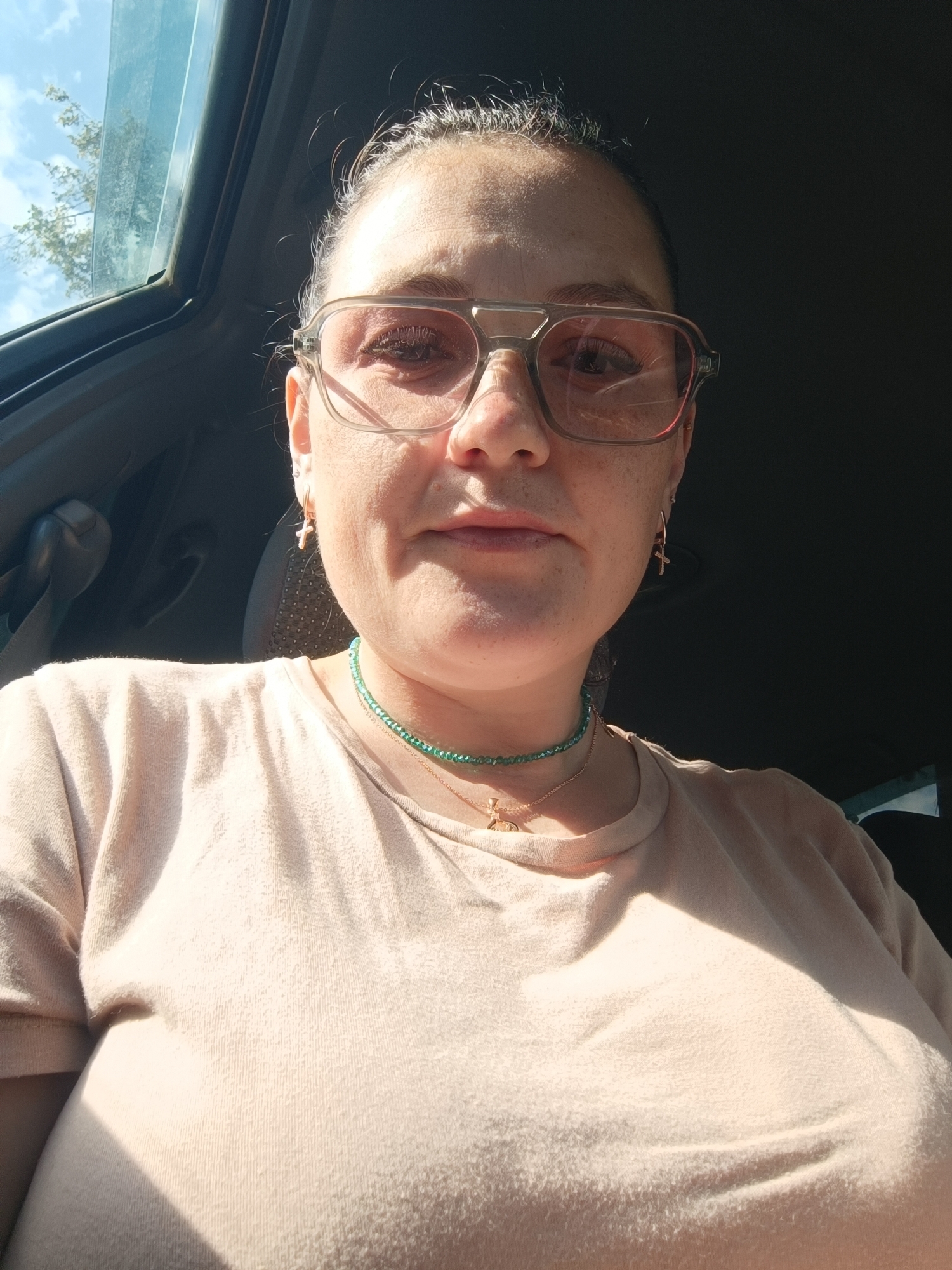 The image is a selfie taken from the chest up of a woman wearing large, light pink-tinted glasses and a light-colored t-shirt. She has dark hair pulled back and is wearing a green beaded necklace with a gold cross pendant and smaller gold cross earrings. The lighting suggests she is indoors or in a shaded area, with sunlight casting shadows across her shirt and face. The background is dark, with a sliver of the sky and tree branches visible through what appears to be a sunroof or window at the top left. No specific location in Chaikovsky, Russia is discernible from the image.