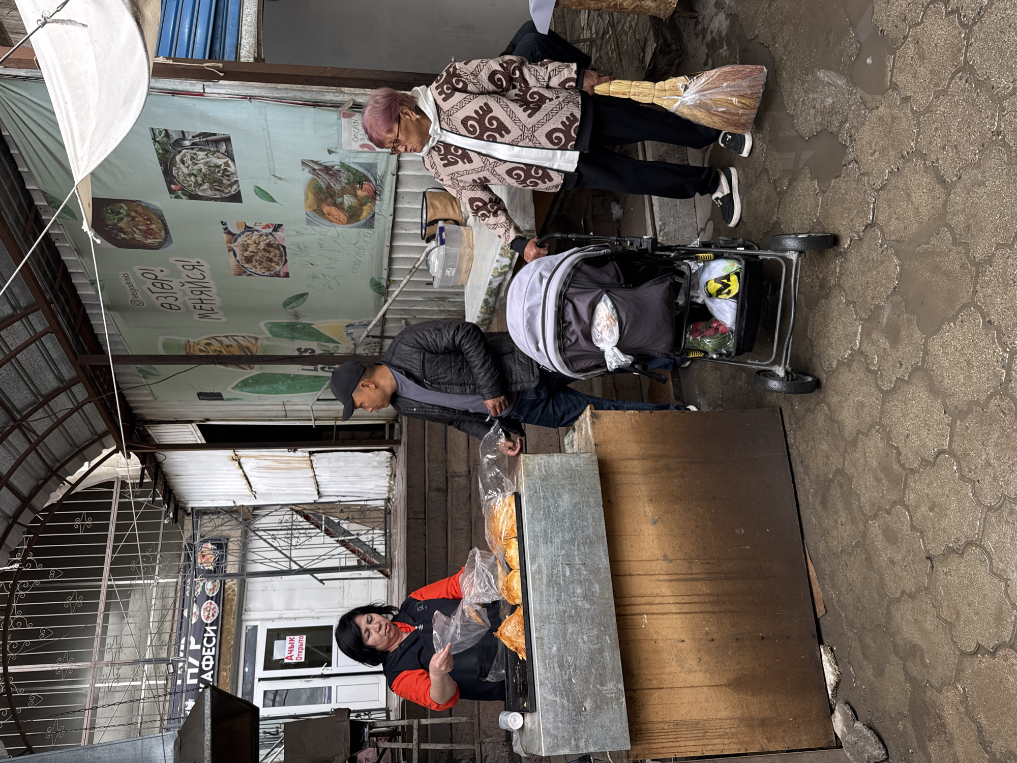 This image captures a bustling street market scene in Bishkek City, Kyrgyzstan, under what appears to be an overcast sky, with the wet ground and puddles suggesting recent rain.
In the foreground, a woman with dark hair, wearing a black vest over an orange shirt, is actively working behind a metal display counter. The counter is filled with various baked goods, possibly bread or pastries, some of which are already packaged in clear plastic bags. She appears to be engaged in the process of selling or preparing items.
Adjacent to the sales stall, a man wearing a black baseball cap and jacket stands next to a baby stroller. The stroller, with its light-colored canopy, contains several items, including a bright yellow bag and a red plastic bag. Nearby, an older woman with distinctive purple-tinted hair, glasses, and a patterned brown and white geometric jacket holds a bundle of brooms. She is looking down, seemingly interacting with the man or the stroller.
The overhead structure features a simple white canvas awning and a metal framework. In the background, banners and signs add to the market's ambiance. A large green banner displays images of various food dishes, such as noodles, soup, and rice, alongside text including "letsgodrinks," "өзгер! МЕНЯЙСЯ!" (meaning "Change! Change!" in Kyrgyz/Russian), "S", "кола" (cola), and "CHICKEN". Another vertical banner prominently displays "КАФЕСИ" (CAFE). A nearby door features signs in both Kyrgyz ("Ачык") and Russian ("Открыто"), both indicating "Open." The overall scene depicts a slice of daily life with vendors and customers in a local market setting.
