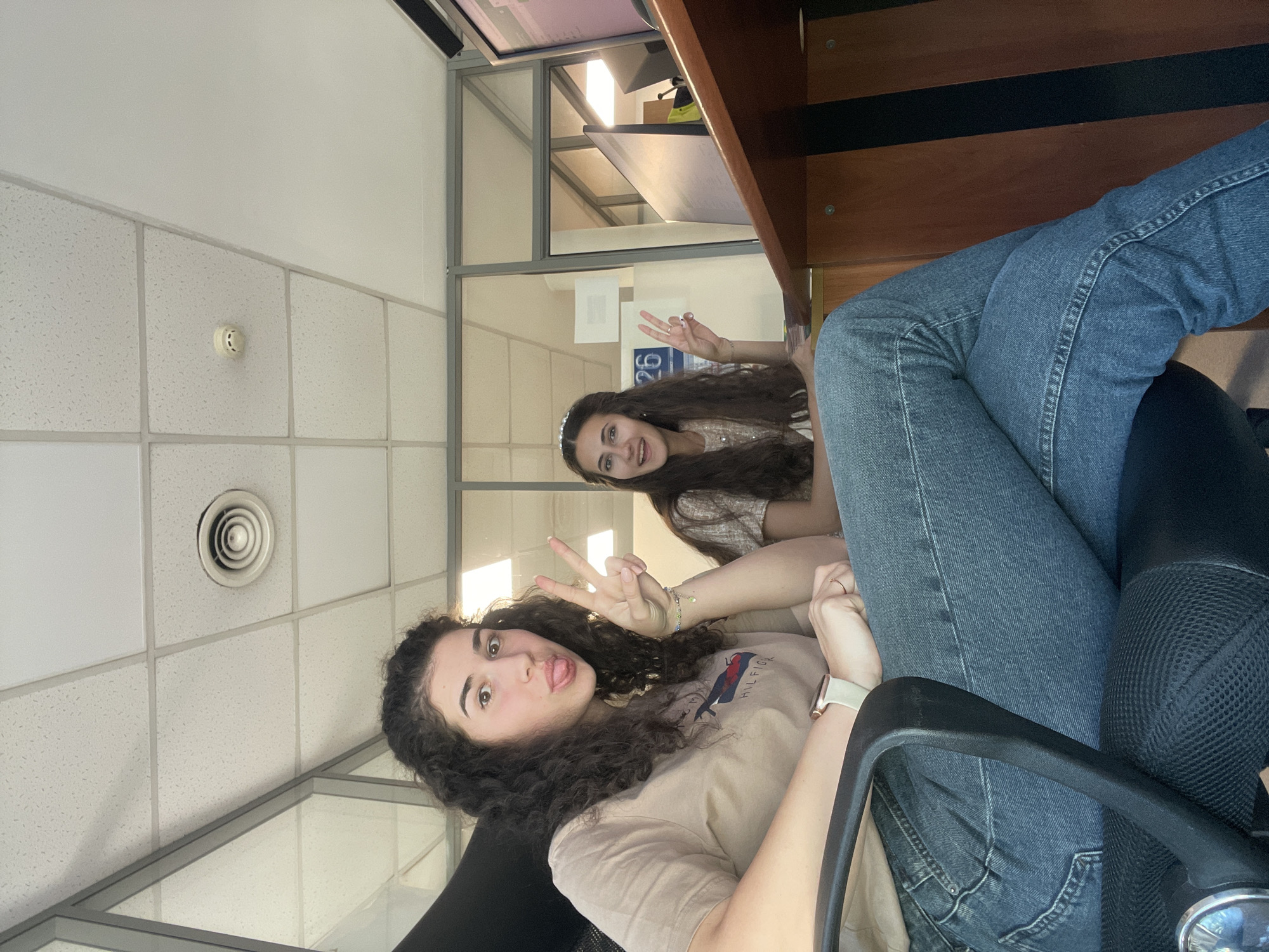 Two young women are posing in what appears to be an office setting. The first woman, in the foreground, has curly dark hair, wears a beige shirt, a watch on her wrist, and makes a peace sign with her fingers while sticking out her tongue. The second woman, in the background, also has long dark hair, wears a light-colored top, and holds up a peace sign. The office has tiled ceilings, ceiling vents, and a fire alarm. There is a desk with a computer monitor. A paper with the number "26" is visible in the background. The location is likely an office interior but a specific location in Sofia, Bulgaria, cannot be determined.