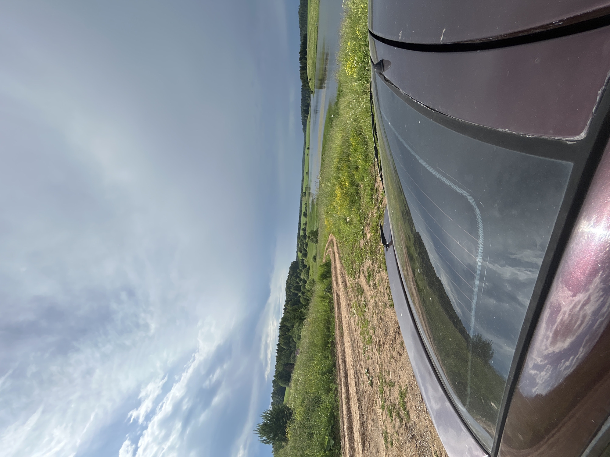 The image captures a rural landscape near Verkh-Rozhdestvo, Russia. In the foreground, the dark purple roof and rear window of a car are visible, angled towards the right. The rear window reflects the sky and surrounding greenery. A dirt road curves through a grassy field dotted with yellow wildflowers, leading towards a body of water. The water, likely a river or lake, stretches into the distance, bordered by green fields and a dense line of trees on the horizon. The sky is dramatic, with a mix of bright clouds and darker, stormier formations overhead. The overall scene conveys a sense of natural beauty and a journey through the Russian countryside.