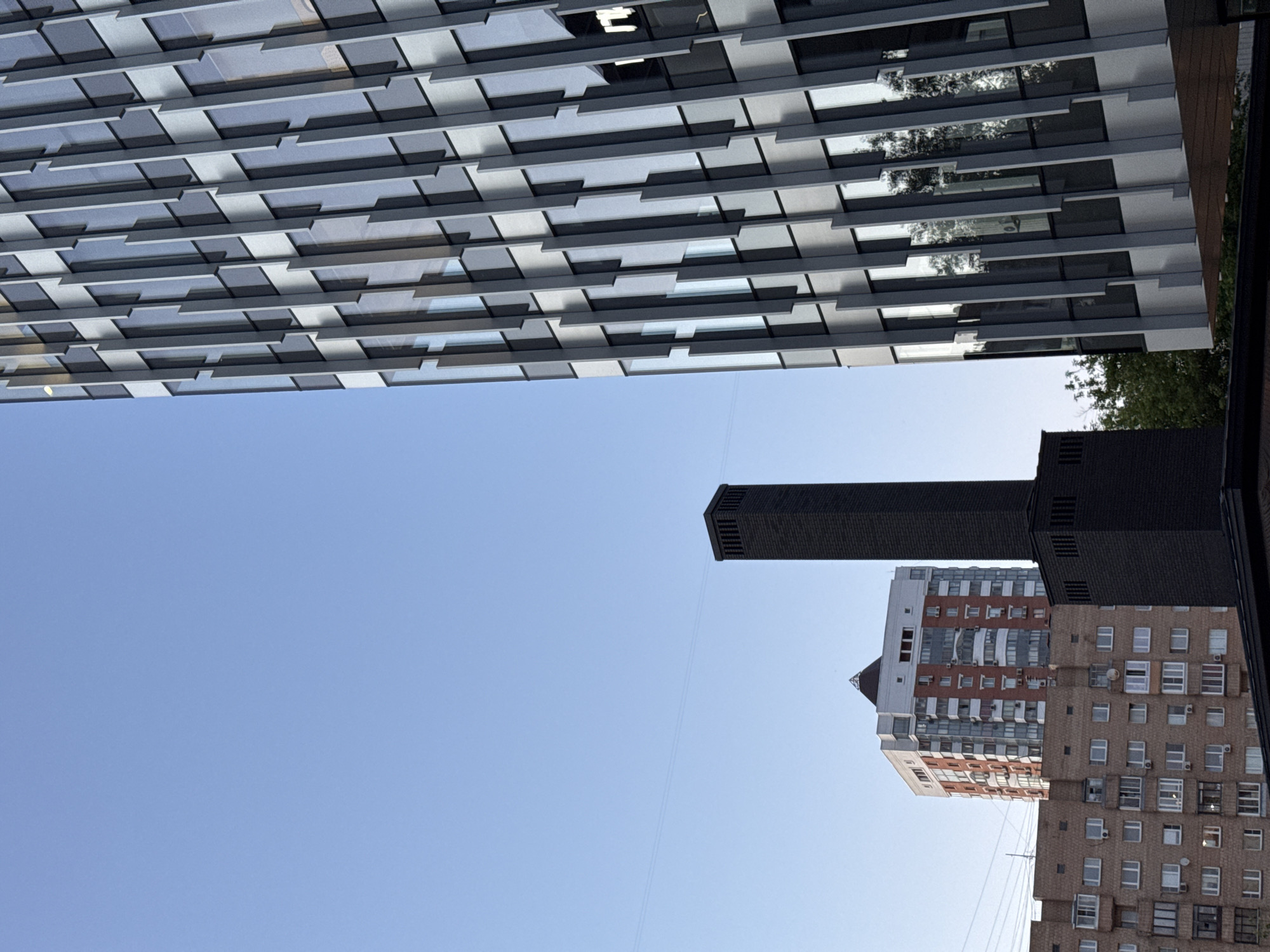 The image depicts an urban landscape in Moscow, Russia, under a clear, light blue daytime sky. On the right, a modern high-rise building with a distinctive facade occupies a significant portion of the frame. This facade is composed of repetitive vertical elements, alternating between reflective glass panels and matte grey or silver architectural fins, creating a dynamic, textured surface. Reflections of green foliage and the sky are visible in the glass sections.
Centrally positioned, a tall, slender, dark-colored rectangular chimney or ventilation shaft stands prominently. It is constructed from dark brick or similar material, featuring horizontal vents near its apex and a slightly wider base.
Behind the dark shaft and to the left, a multi-story residential building is visible, characterized by a facade that mixes red-brown brick sections with lighter, possibly white or cream-colored, panels. This building displays numerous windows and multiple exterior air conditioning units, and its roof is topped with a small, pointed, pyramidal structure.
In the lower-left quadrant, an older, multi-story residential building, constructed of brown-toned brick, is partially visible, showing multiple windows and balconies. Green tree foliage is present behind the dark shaft and reflected in the modern building's windows. No individuals, vehicles, or specific activities are discernible in the scene.