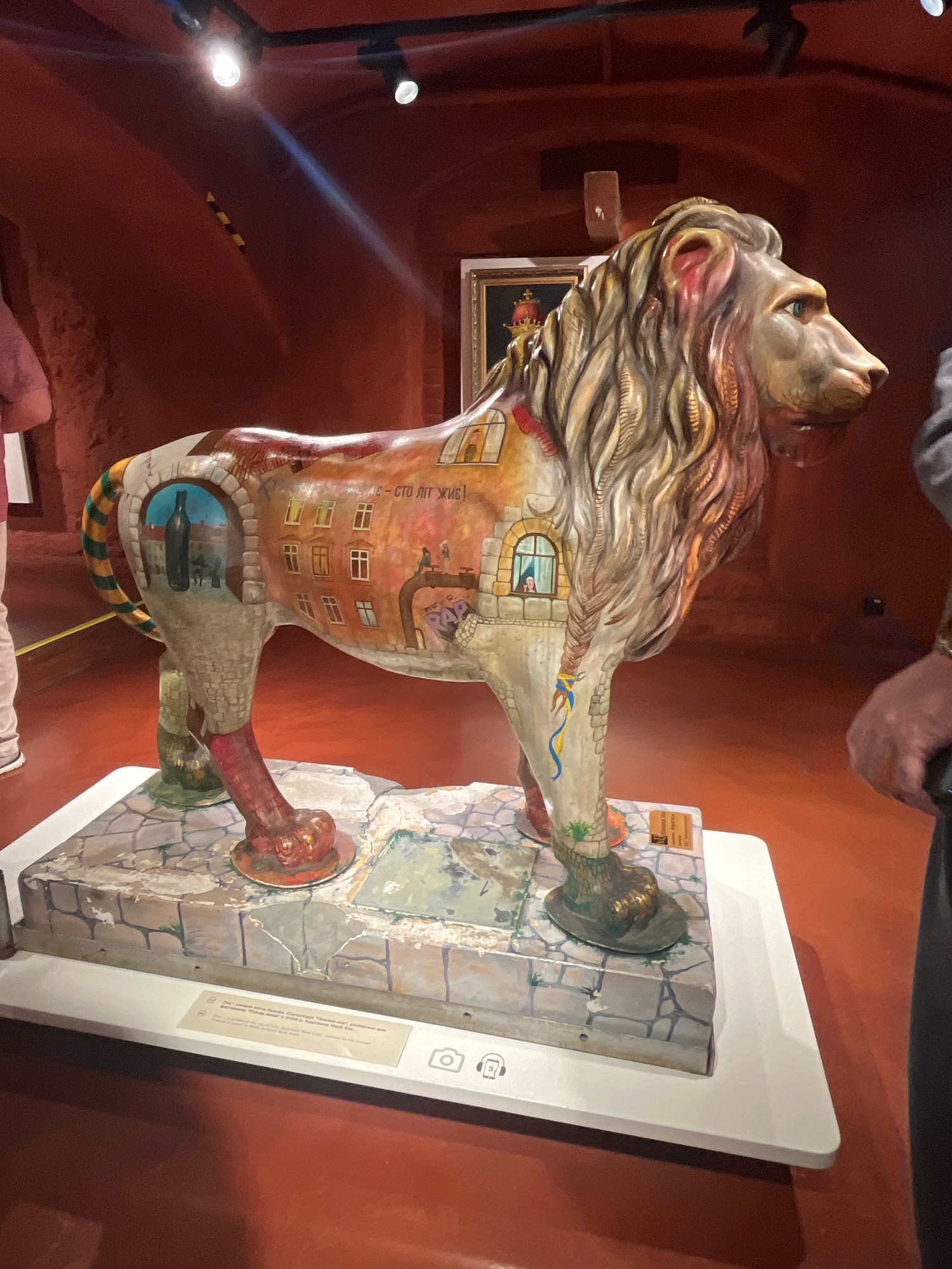 This image shows a brightly painted statue of a lion displayed on a pedestal in what appears to be a museum or gallery. The lion sculpture is decorated with detailed scenes of urban life, including buildings, windows, and people. The background is a deep red, suggesting a curated exhibition space. The sculpture is part of an art installation called "Lions of Lviv," which is a city-wide project in Lviv, Ukraine, featuring numerous lion sculptures decorated by various artists. These sculptures are placed throughout the city, often in public spaces, as a way to celebrate the city's identity and history, as the lion is a prominent symbol of Lviv. The statue in the image is one of these numerous art pieces, contributing to the unique visual character of Lviv.