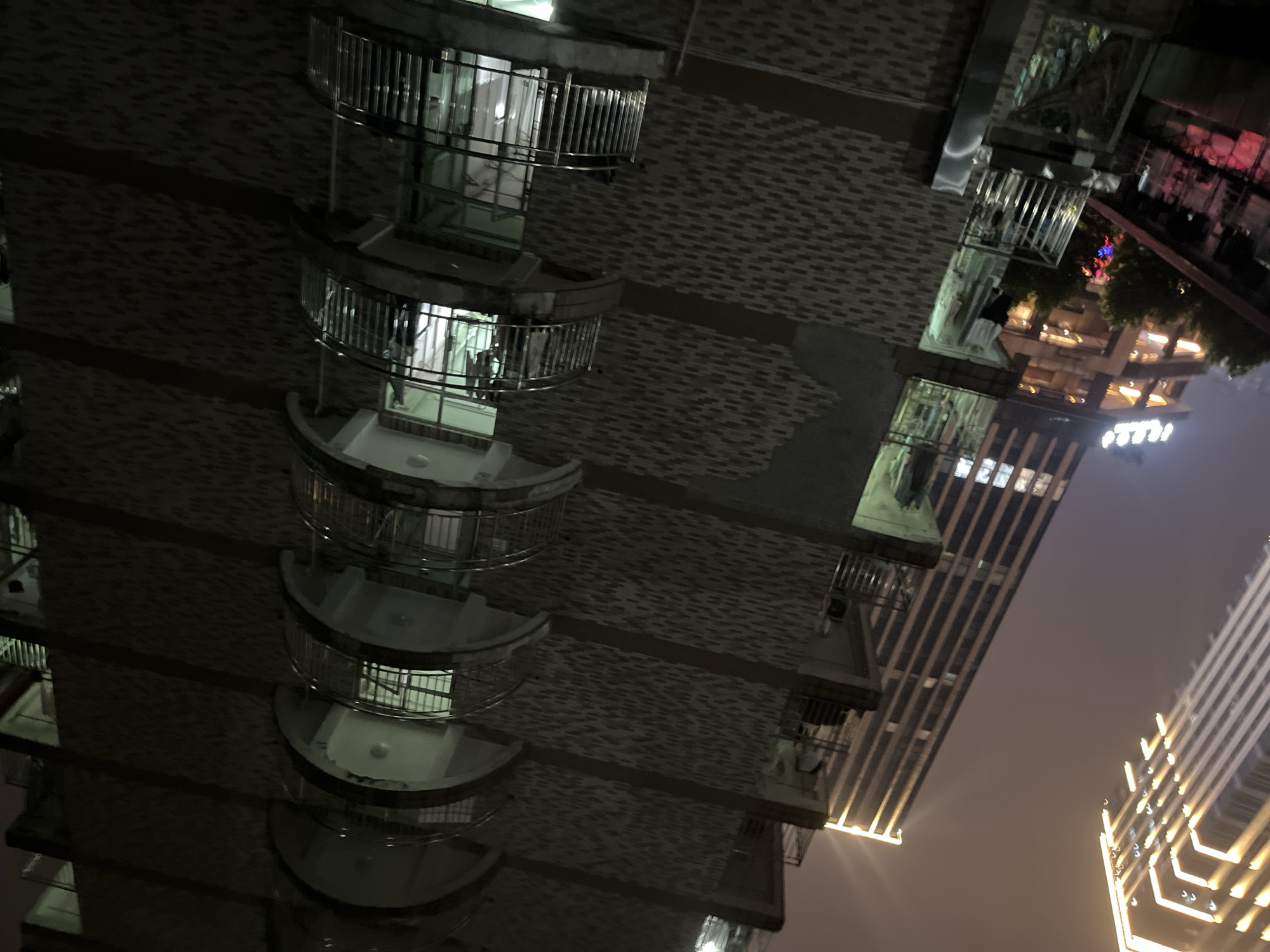 This image shows a night view of a multi-story residential building with balconies, located in Yuzhong District, China. The building's facade is constructed with a textured brick pattern. Each balcony features a curved design and is enclosed by a silver metal railing. Interior lights are visible in some of the balconies and windows, illuminating the scene with a greenish-white glow. In the background, a taller, modern skyscraper with illuminated vertical accents is visible, suggesting an urban environment. The sky is hazy with a warm, diffused light, possibly from city lights or fog. The perspective is from a lower vantage point, looking up at the buildings.