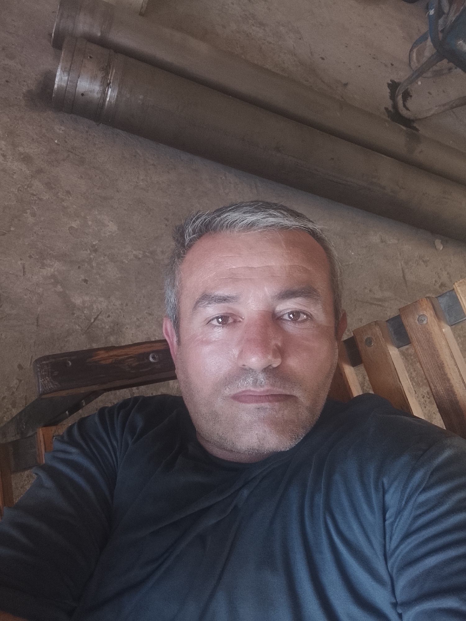 A close-up, selfie-style medium shot shows a middle-aged man with short, graying hair and light stubble. He is wearing a dark, long-sleeved t-shirt and is seated in front of a rough, concrete wall. To his left, a wooden chair with metal fasteners is visible. In the background, two large, cylindrical metal pipes are stacked horizontally against the wall. The lighting suggests an indoor setting, possibly a workshop or storage area, with a shadow cast on the wall and pipes from an unseen light source. The image is taken from a slightly upward angle, focusing on the man's face and upper torso.