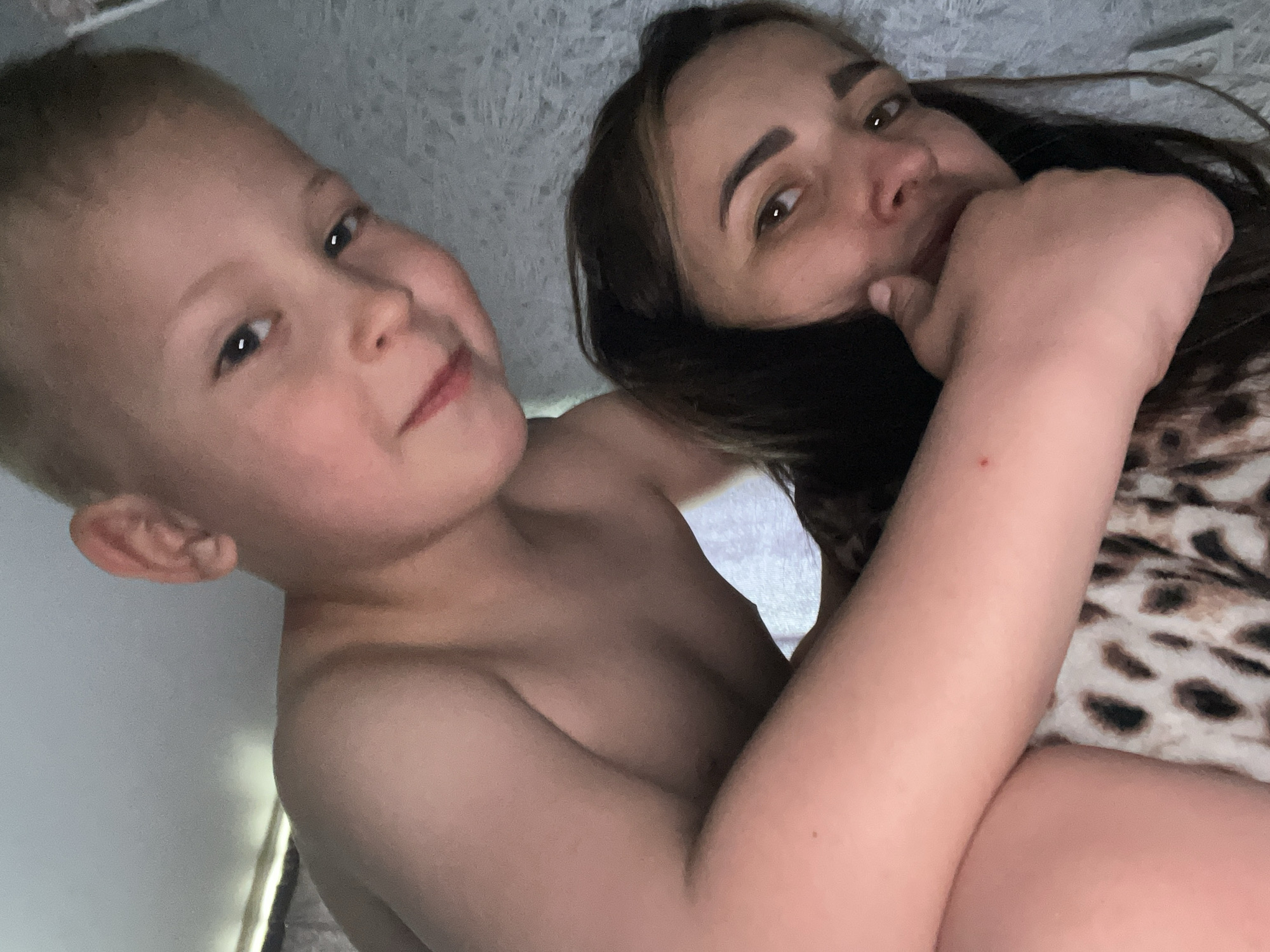 A light-skinned woman with dark hair and a fair-skinned, young, blond boy are pictured from a close, upward-facing angle. The boy is shirtless and faces the camera with a slight smile. The woman is lying on a leopard print blanket, with her hand near her face. The background consists of a white textured surface that appears to be a wall or ceiling. The location is Unknown, Unknown.