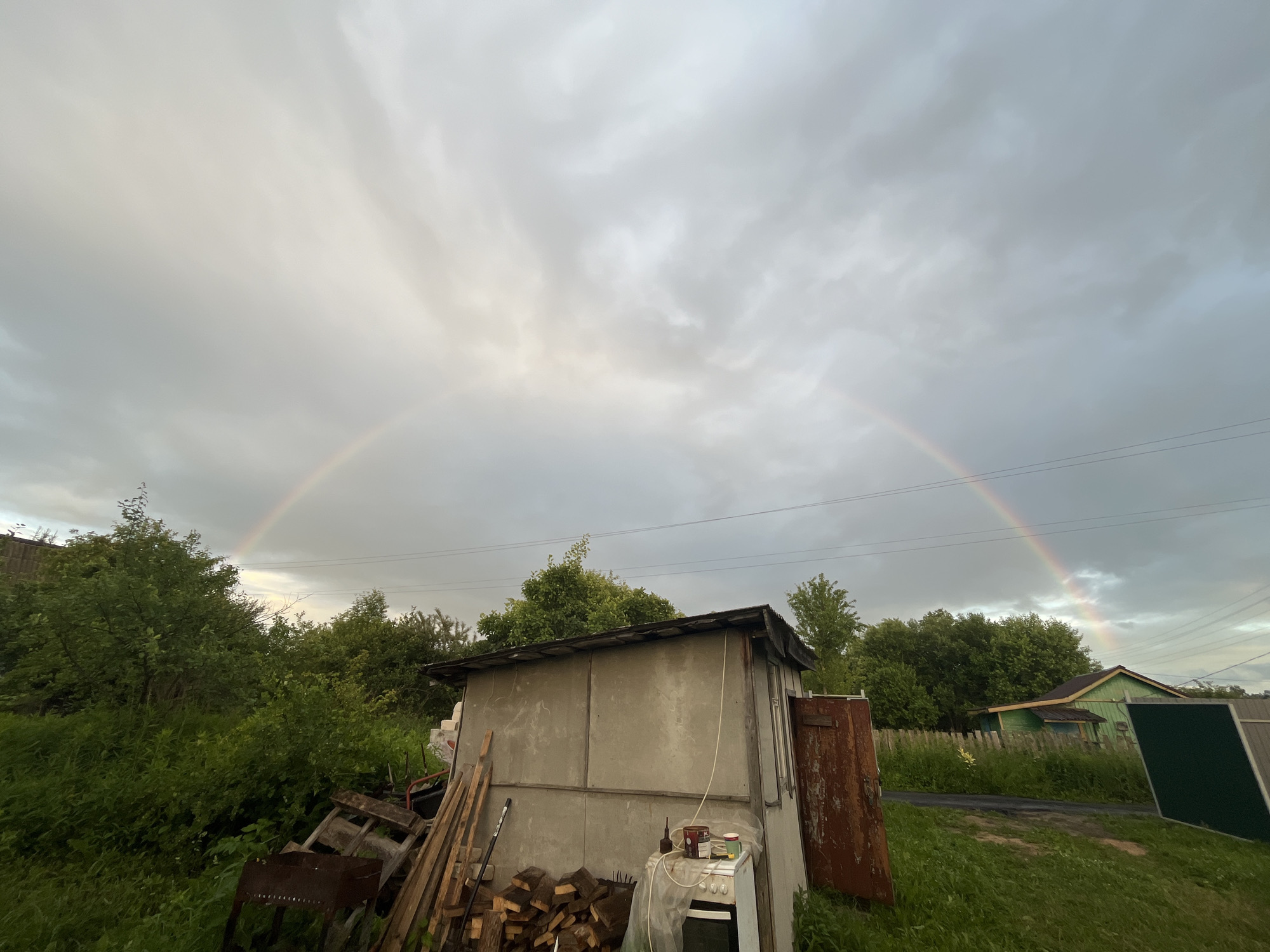 The image captures a cloudy sky with a faint double rainbow stretching across it, located in the village of Klobushnevo, Russia. In the foreground, a dilapidated concrete shed stands to the right, with a rusty metal door and various discarded items around it, including stacked firewood, wooden planks, and a rusty barbecue grill. Lush green foliage and trees surround the shed and extend into the background, where a small green house with a dark roof is visible. Power lines are strung across the sky above the trees. The overall scene suggests a rural, perhaps slightly neglected, outdoor setting after a rain shower.