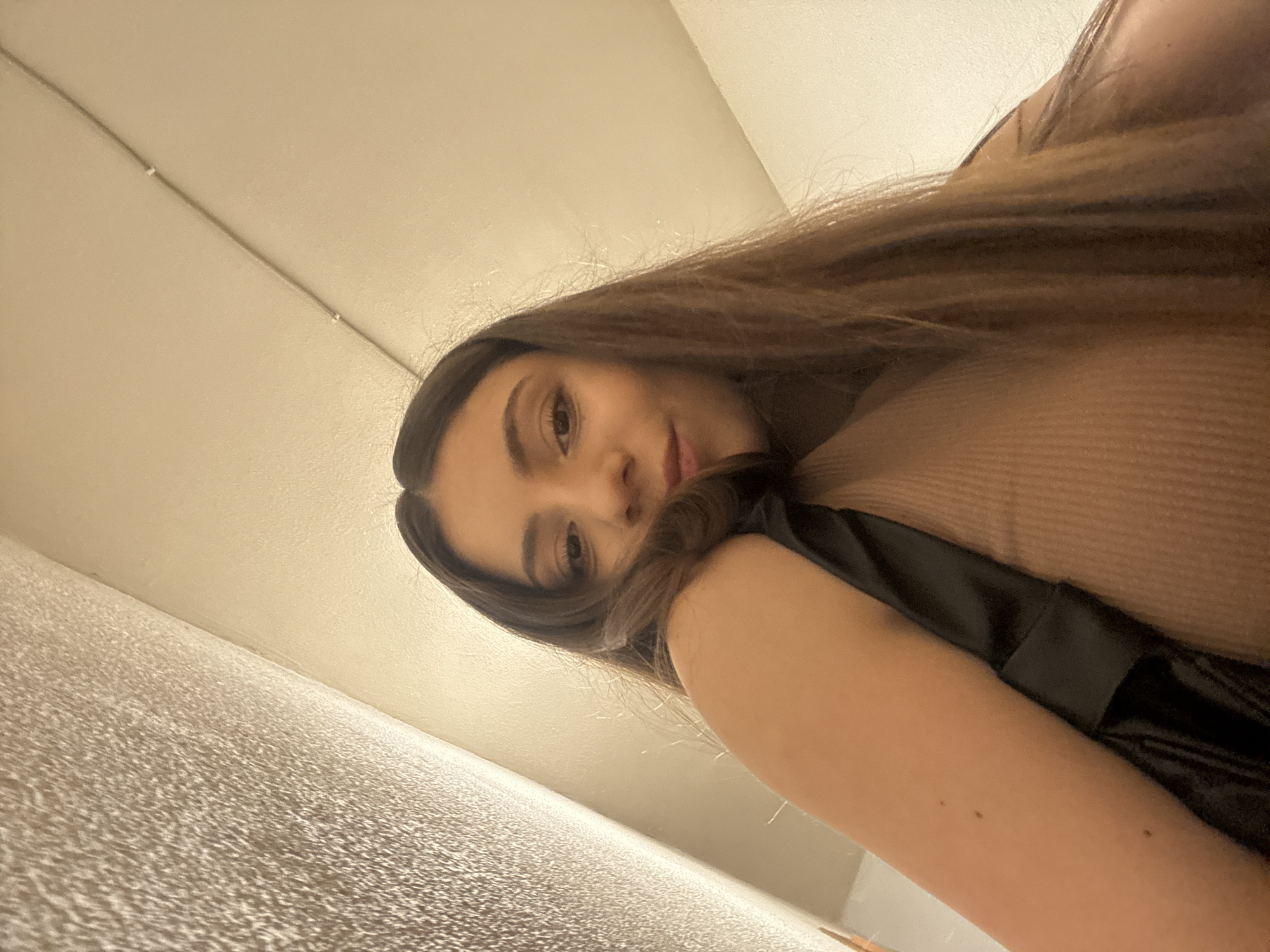 The media file is a first-person perspective, low-angle selfie of a young woman. She has long, dark brown hair that falls over her face and shoulder. Her face is partially obscured, but her eyes are visible, and she appears to be looking directly at the camera with a soft smile. She is wearing a light brown, ribbed garment and a black strap or garment is visible across her arm and chest. The background consists of a textured, light-colored surface that appears to be a wall, and a pale ceiling. A thin, dark wire runs diagonally across the upper portion of the frame. The lighting suggests an indoor setting, possibly during the daytime or with artificial light. There is no visible text in the image. The overall mood is casual and intimate, typical of a personal selfie.