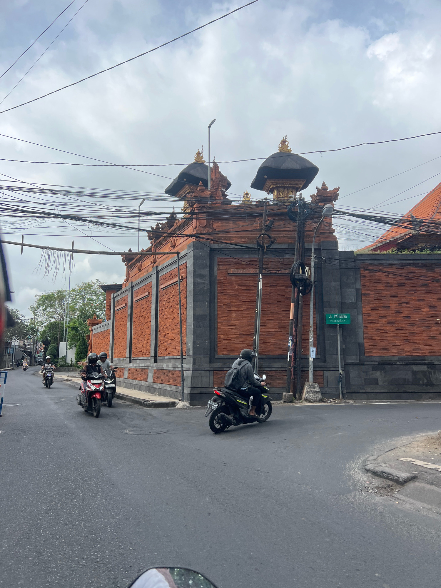 The image depicts a street scene in Kuta, Indonesia, featuring a large, ornate brick building with a dark, tiered roof and gold accents. A green street sign indicates "JL. PATIMURA". Several individuals are riding motorbikes on the paved road. Two motorbikes are in the foreground, with riders wearing helmets. Another motorbike is further down the road, also with a rider. Overhead, a complex network of electrical wires crisscrosses the sky, obscuring parts of the cloudy sky. The building appears to be a temple or a similar cultural structure, characterized by its detailed brickwork and architectural elements. The overall environment suggests a typical urban setting in Indonesia, with motorbikes being a primary mode of transportation.