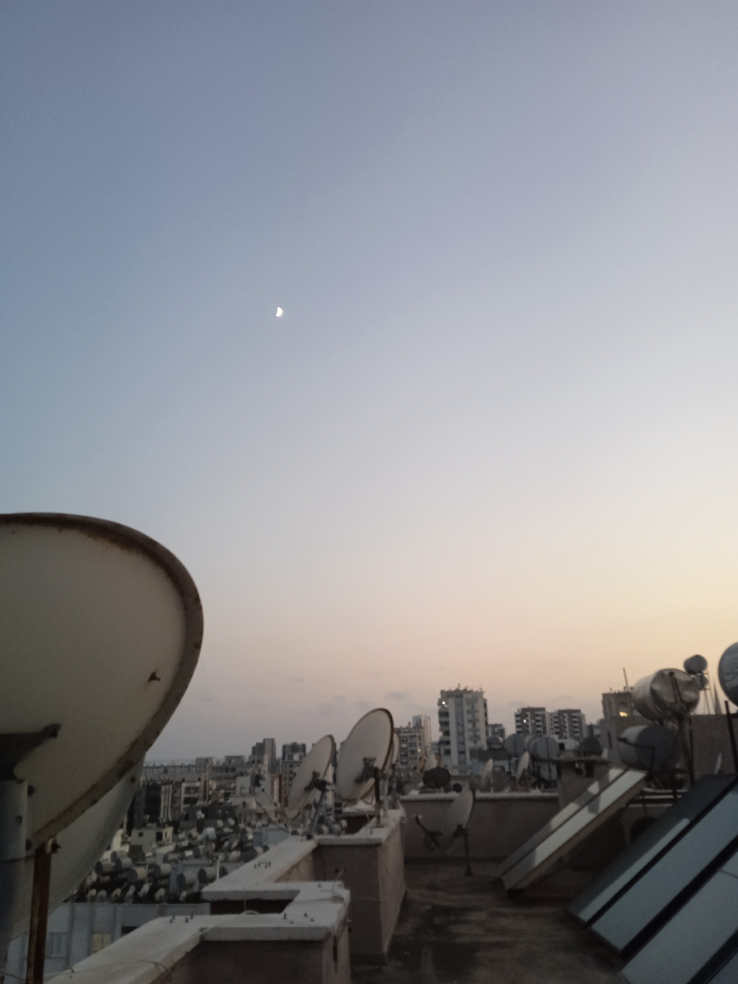 A rooftop scene is depicted at dusk in Mezitli, Turkey, with a crescent moon visible in the pale blue sky. The foreground features a large satellite dish in the lower left corner. Beyond this, a cityscape stretches across the horizon, characterized by numerous apartment buildings and a dense arrangement of satellite dishes and solar water heaters on various rooftops. The overall impression is one of a densely populated urban environment in the early evening.