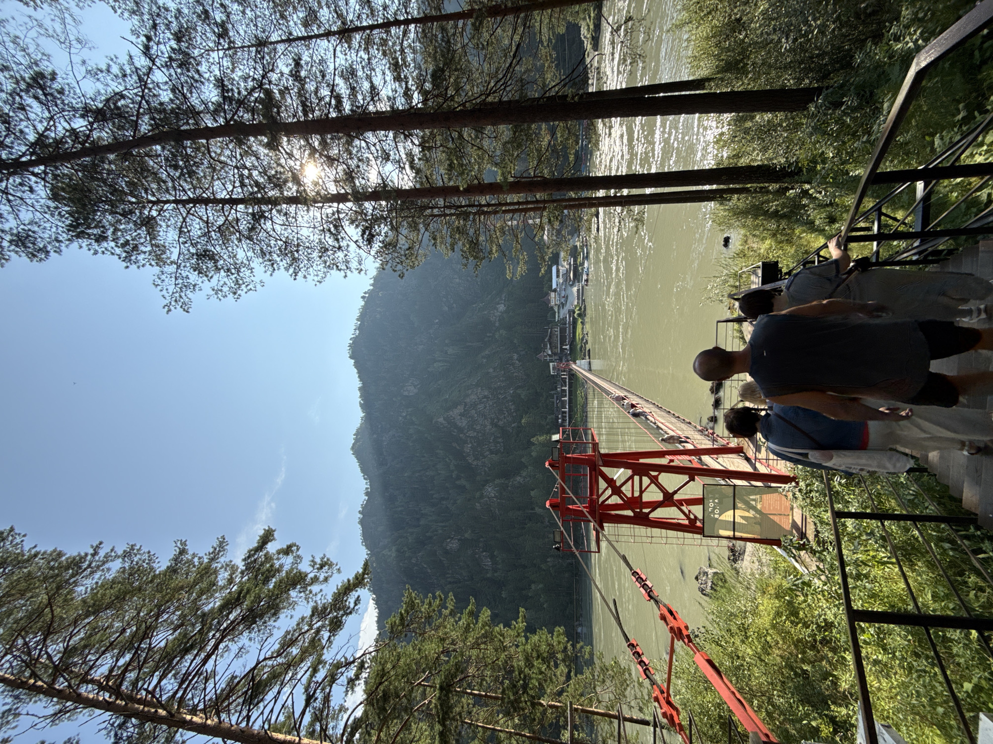 The image captures a sun-drenched, scenic view of a suspension bridge crossing a wide, greenish river in Barangol, Russia. Tall pine trees frame the scene, their branches reaching towards a clear blue sky. The bridge, painted a striking red, features robust support towers and a long walkway populated by a few pedestrians. The river flows with visible ripples, reflecting the sunlight. In the background, a lush, tree-covered mountain range rises majestically, with a cluster of buildings and boats visible on the far bank of the river, indicating a settlement. In the foreground, a group of people are descending a set of stairs leading towards the bridge, their backs to the camera. The overall atmosphere is one of natural beauty and outdoor recreation.