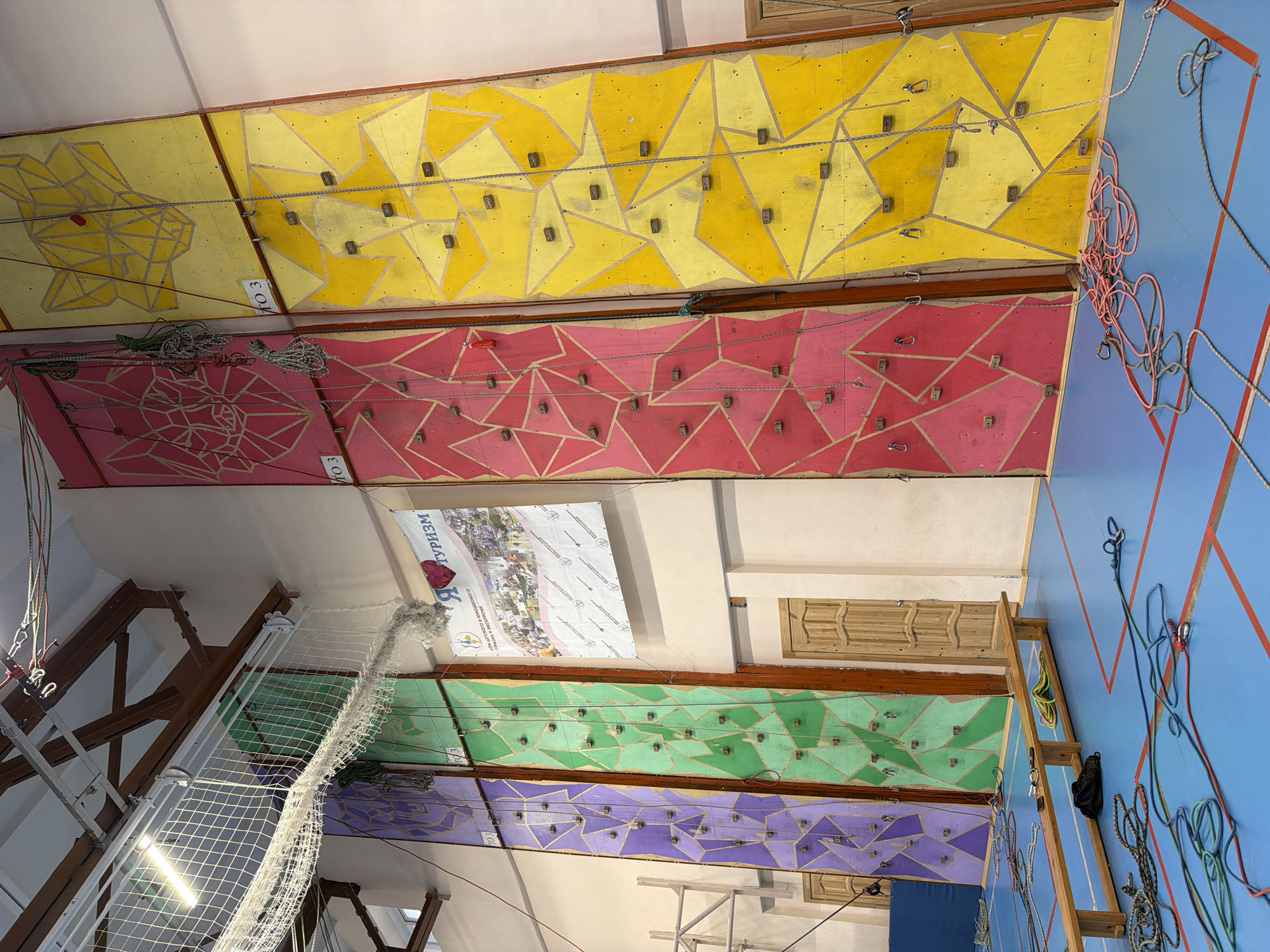 The image depicts an indoor rock climbing facility. The climbing walls are segmented into horizontal bands, each painted a distinct color: yellow, red, green, and purple. Each band features a geometric pattern painted in a lighter shade of the base color. Climbing holds are bolted onto these walls, forming routes. Ropes and carabiners are suspended from the top of the walls. A safety net is visible to the left. A blue wall is located on the right, with red tape creating a triangular design. A wooden ladder leans against this wall. A poster in Russian is affixed to the wall between the red and green climbing bands. Text on the poster mentions "ТУРИЗМ" (Tourism). The location is Berezniki, Russia, based on the banner.