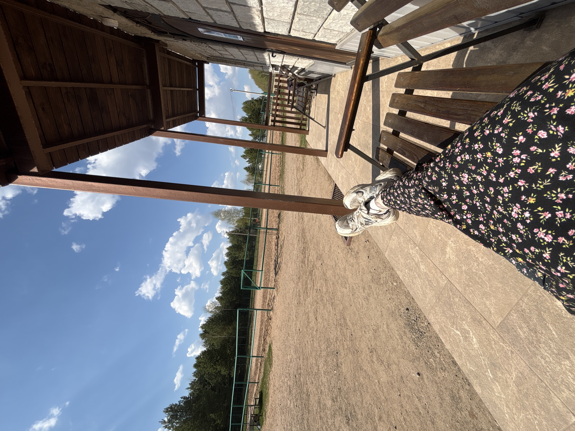 The image is a tilted perspective shot, possibly taken from a low angle. It shows a person sitting on a wooden bench. The person is wearing a black floral dress. Their white sneakers are visible.
The bench is part of a structure that appears to be a porch or veranda. The structure has a wooden roof and is attached to a building with brick or stone exterior walls.
Beyond the structure is an open field. A green fence runs along the field's edge, separating it from a line of trees. The sky is blue with scattered white clouds.
Based on the prompt, the location is Русский Вожой, Russia.