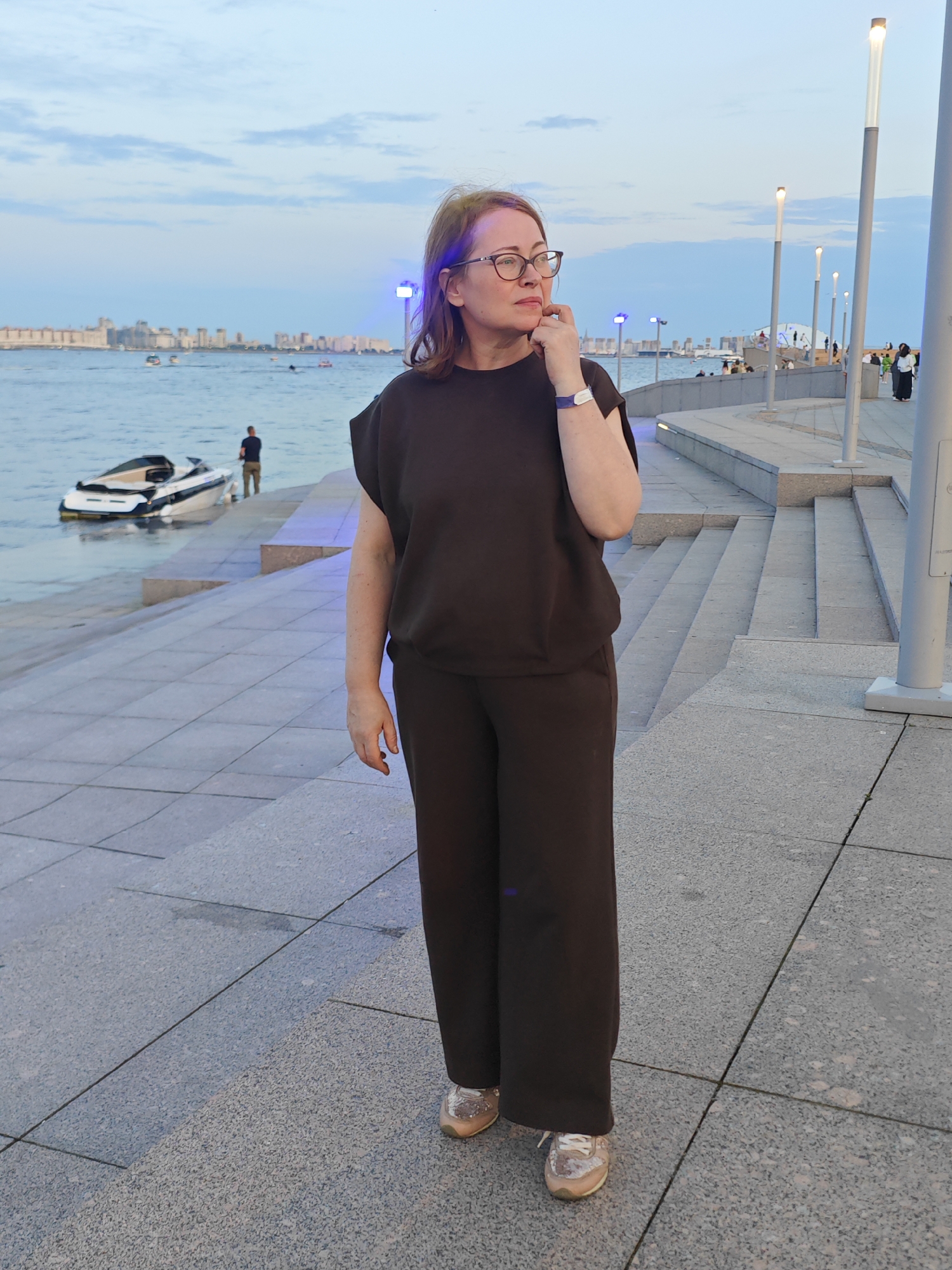 A woman wearing glasses and a dark brown tracksuit stands on a stone-tiled staircase by a body of water in Saint Petersburg, Russia. The woman is looking to her right, with her hand near her chin. In the background, a cityscape with buildings is visible across the water, along with several boats and a jet ski. There are streetlights and other people in the background. The sky is partly cloudy, and the lighting suggests it is dusk.