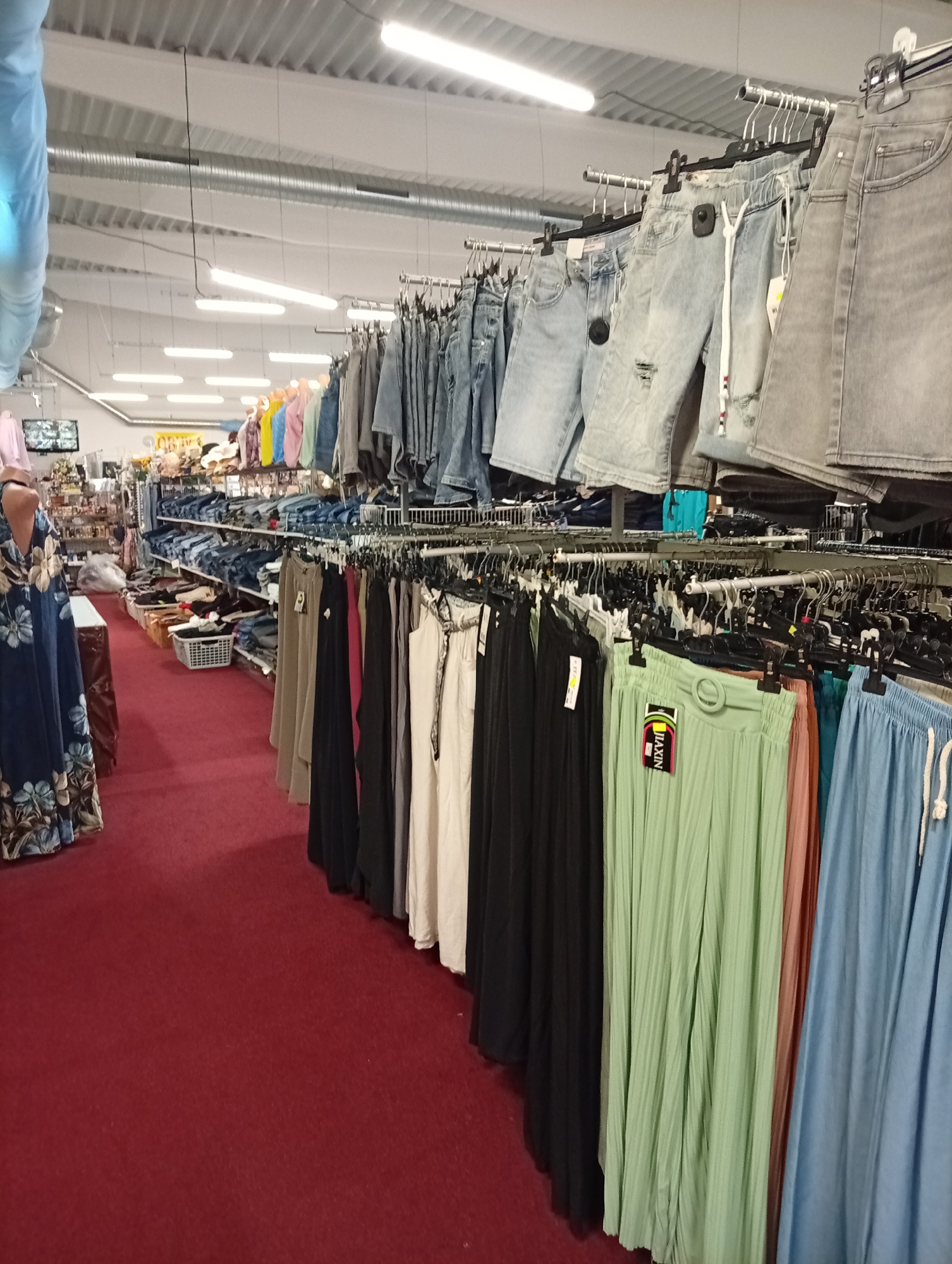 The image shows the interior of a clothing store located in Tišnov, Czechia. The store is well-lit with fluorescent lighting fixtures on the ceiling. The floor is covered with a red carpet, and clothing racks are arranged throughout the space. In the foreground, a variety of pants and shorts are displayed on hangers. These include denim shorts, light blue pleated pants, and black pants. Further into the store, more clothing racks are visible, displaying jeans and other garments. There are also shelves stocked with folded clothing and baskets filled with items. A mannequin dressed in a floral blue dress stands to the left of the frame. The overall impression is that of a busy retail environment offering a wide selection of apparel.