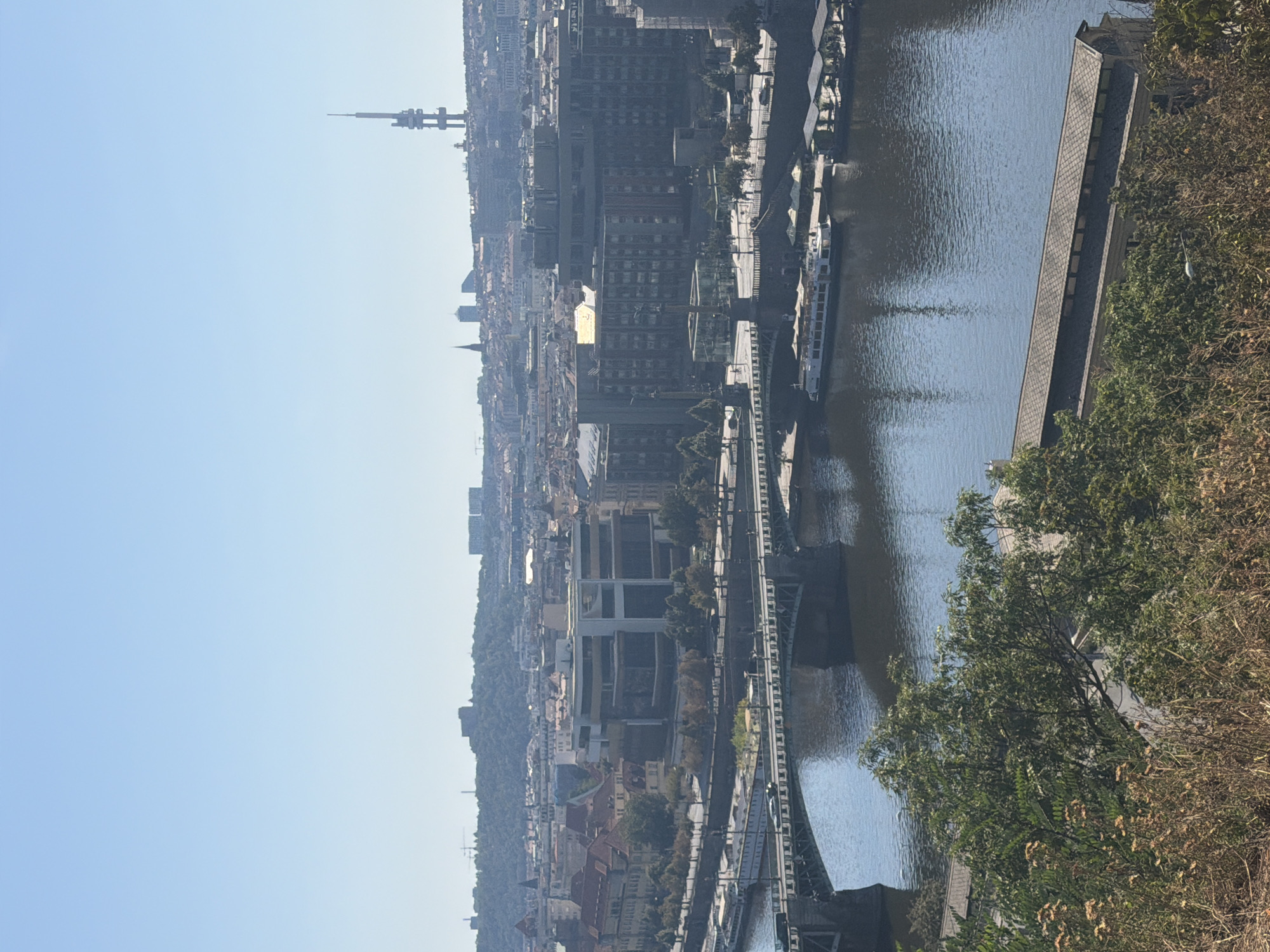The image shows an aerial view of a city, identified as Prague, Czechia. In the foreground, there is dry grass and foliage. A wide river, likely the Vltava River, occupies the middle ground, with a bridge crossing it. Several boats are moored along the riverbanks. On the far bank, a dense urban landscape is visible, characterized by a mix of old and modern buildings, including a prominent broadcast tower in the distance. The sky is clear and blue.
