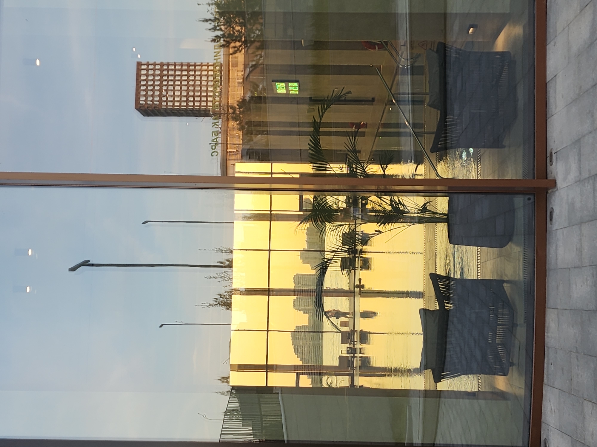 A glass window reflects a building and water in Дачное, Russia. The window frame is brown and the reflection includes modern buildings, trees, and waterfront furniture. The lighting suggests either a sunrise or sunset.