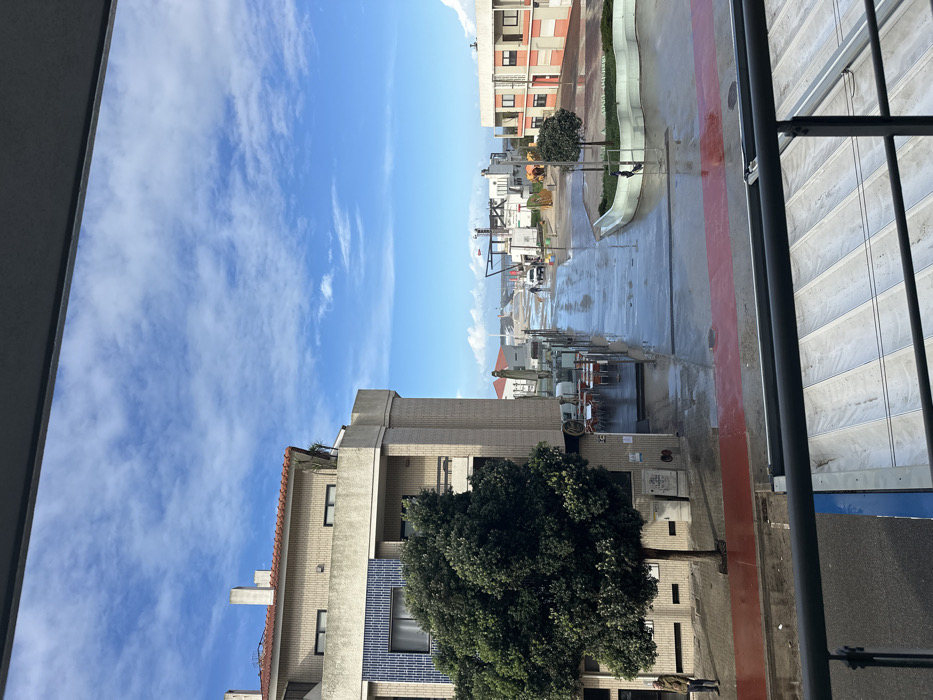 The image displays an elevated view of a wet street and coastal area in Gafanha da Nazaré, Portugal, under a clear blue sky with scattered white clouds. A dark metal balcony railing occupies the bottom fifth of the frame, with a partially visible blue and white striped surface beneath it.
On the left, a multi-story building with a red-tiled roof, beige brick facade, and a distinct blue-tiled section on its lower level stands prominently. A large, dense green tree with a visible trunk is positioned directly in front of this building. Adjacent to the building, a street-level outdoor dining area features several small tables and chairs. Above this area, a life-sized statue, appearing to depict a standing figure in a hat and coat (likely a fisherman), faces towards the distant water. A single individual wearing a light jacket is visible standing on a red-paved path next to the large tree on the far left.
The wide, paved street, visibly wet and reflective, extends from the immediate foreground towards the horizon. A red-paved pedestrian path runs parallel to the street on the left. Further down the street on the right, a smaller multi-story building with orange and beige sections is visible. In front of it, a curved planter contains a small, manicured tree. A second individual is present near this small tree. A white vehicle is discernible further down the wet street. In the background, the street transitions into a sandy shoreline bordering a body of water, presumably the Atlantic Ocean or a connected waterway. Several indistinct structures, consistent with port or beach infrastructure, are visible along the distant coastline and extending into the water. The environment appears to be a coastal promenade or street leading to a beach or harbor.